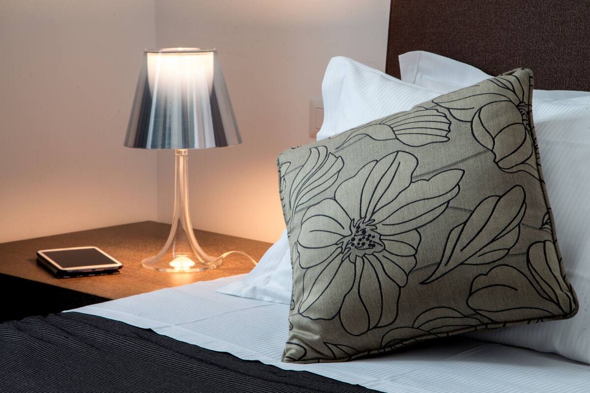 A neatly made bed with white linens, a floral-patterned pillow, a bedside lamp, and a tablet on a nightstand.