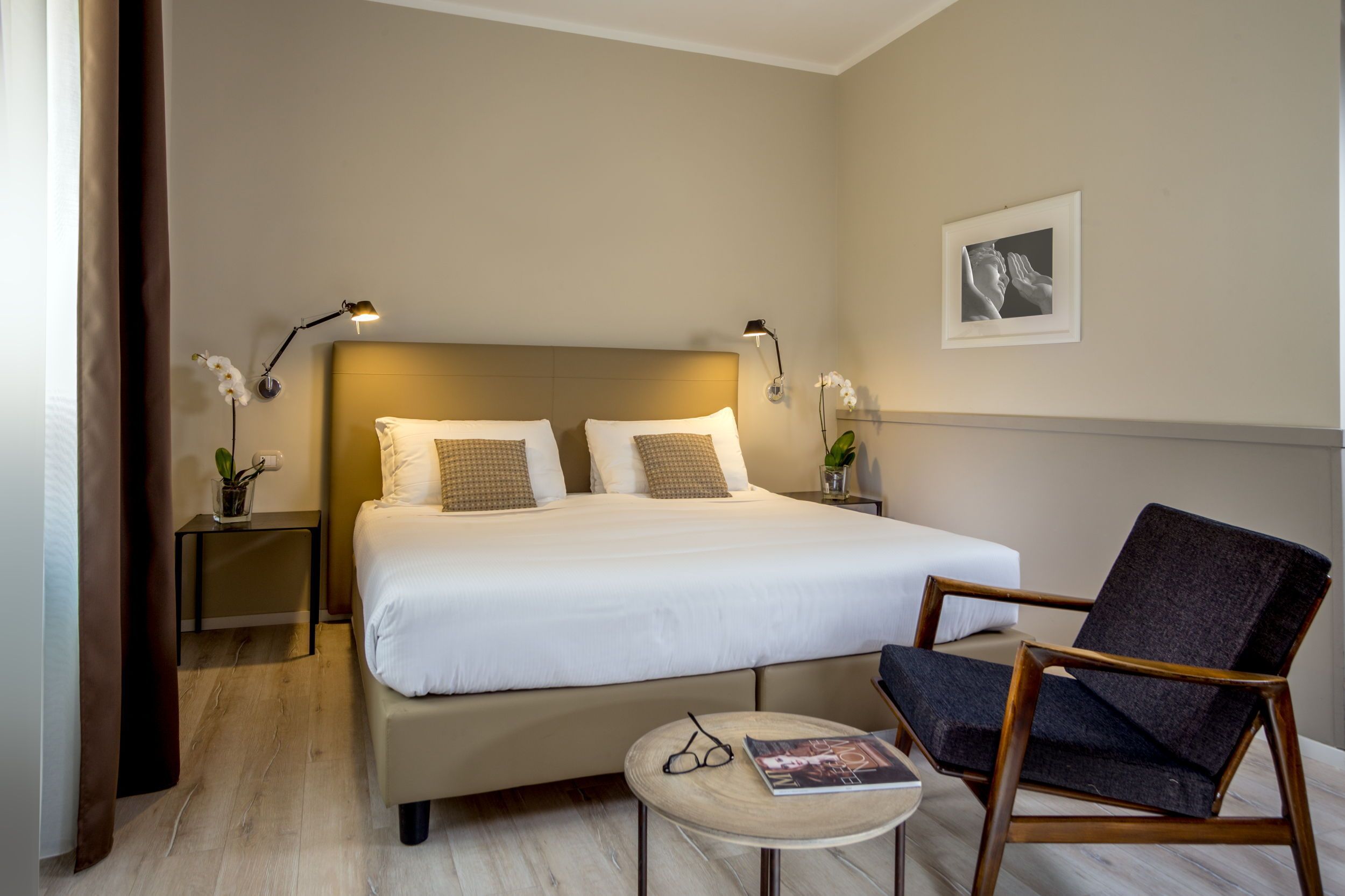 A hotel room with a large bed, two bedside tables with lamps, a chair, a small round table with a magazine, and a framed picture on the wall.