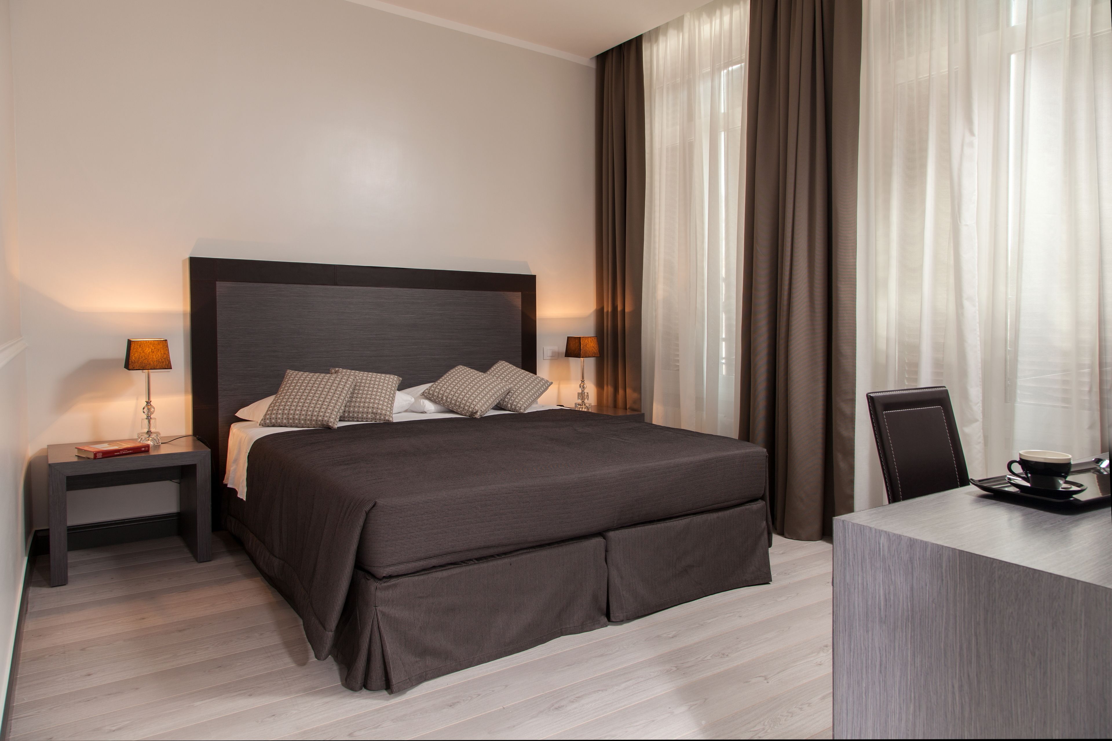 Una habitación de hotel moderna con una cama grande, dos mesitas de noche con lámparas, un escritorio con silla y cortinas translúcidas.