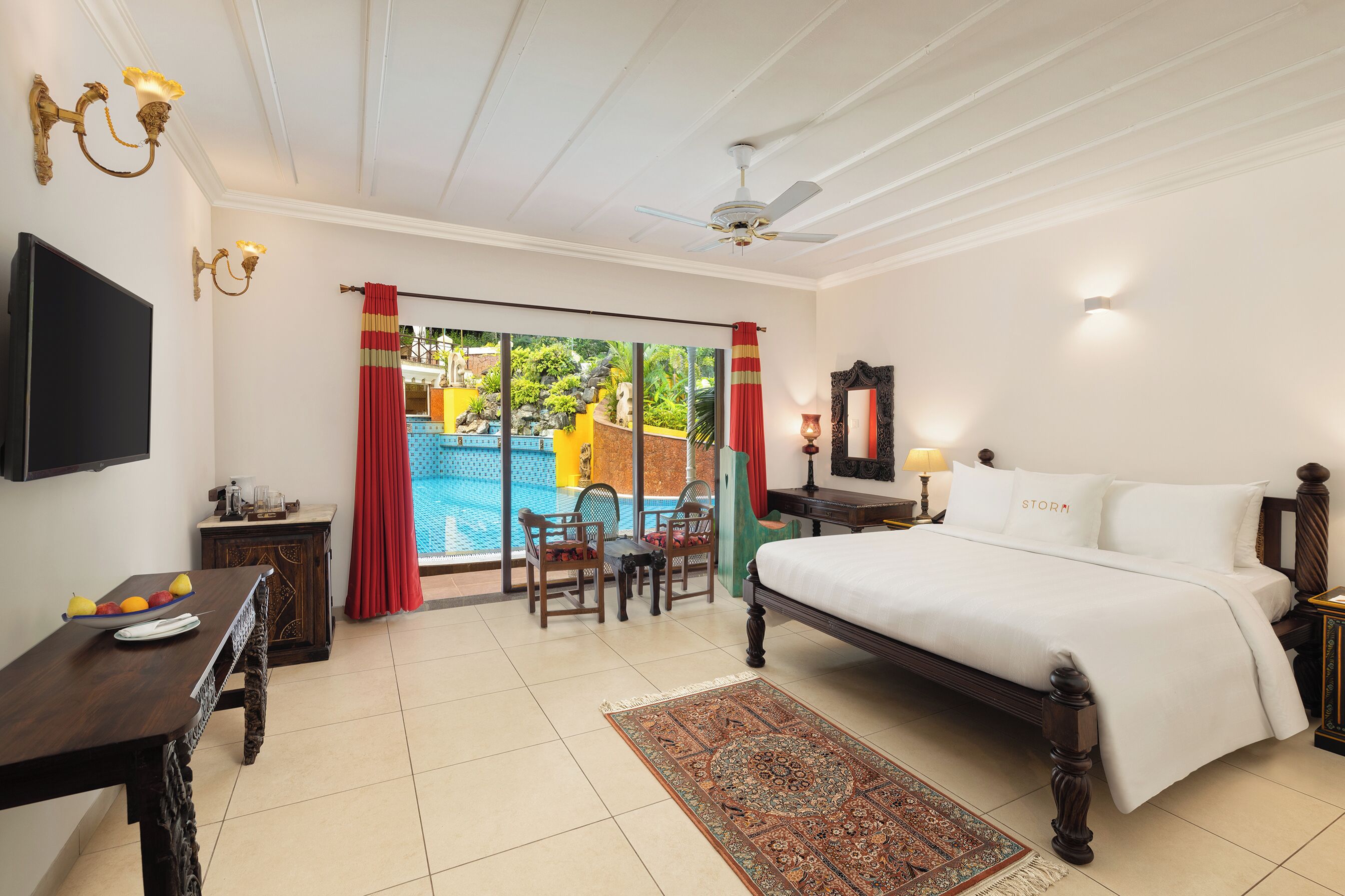 A bedroom with a bed, a desk with a laptop, a TV, a ceiling fan, a window with curtains, and a view of a pool.