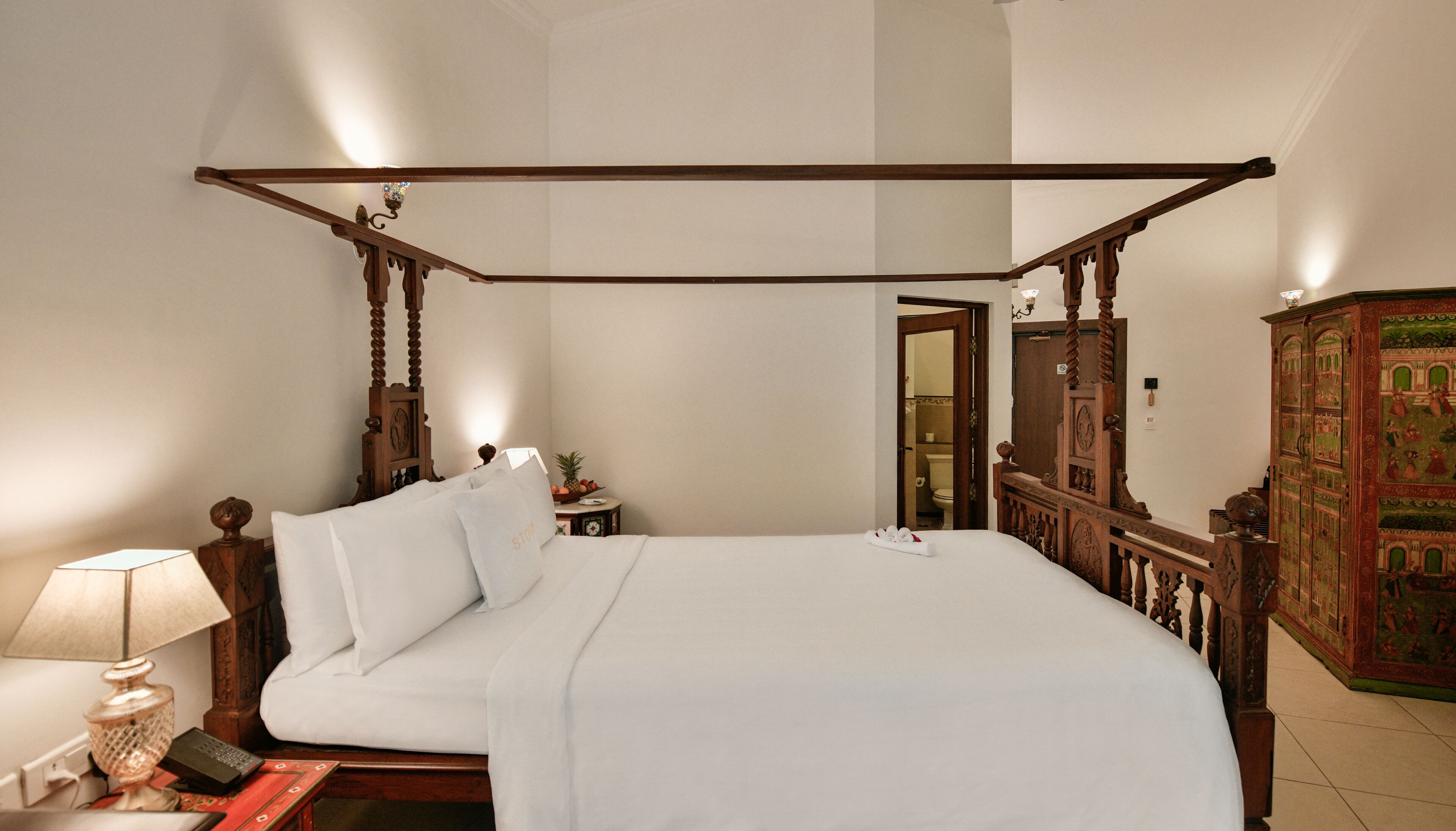 Una cama con dosel y tablero de madera, una mesita de noche con lámpara y un mueble decorativo.