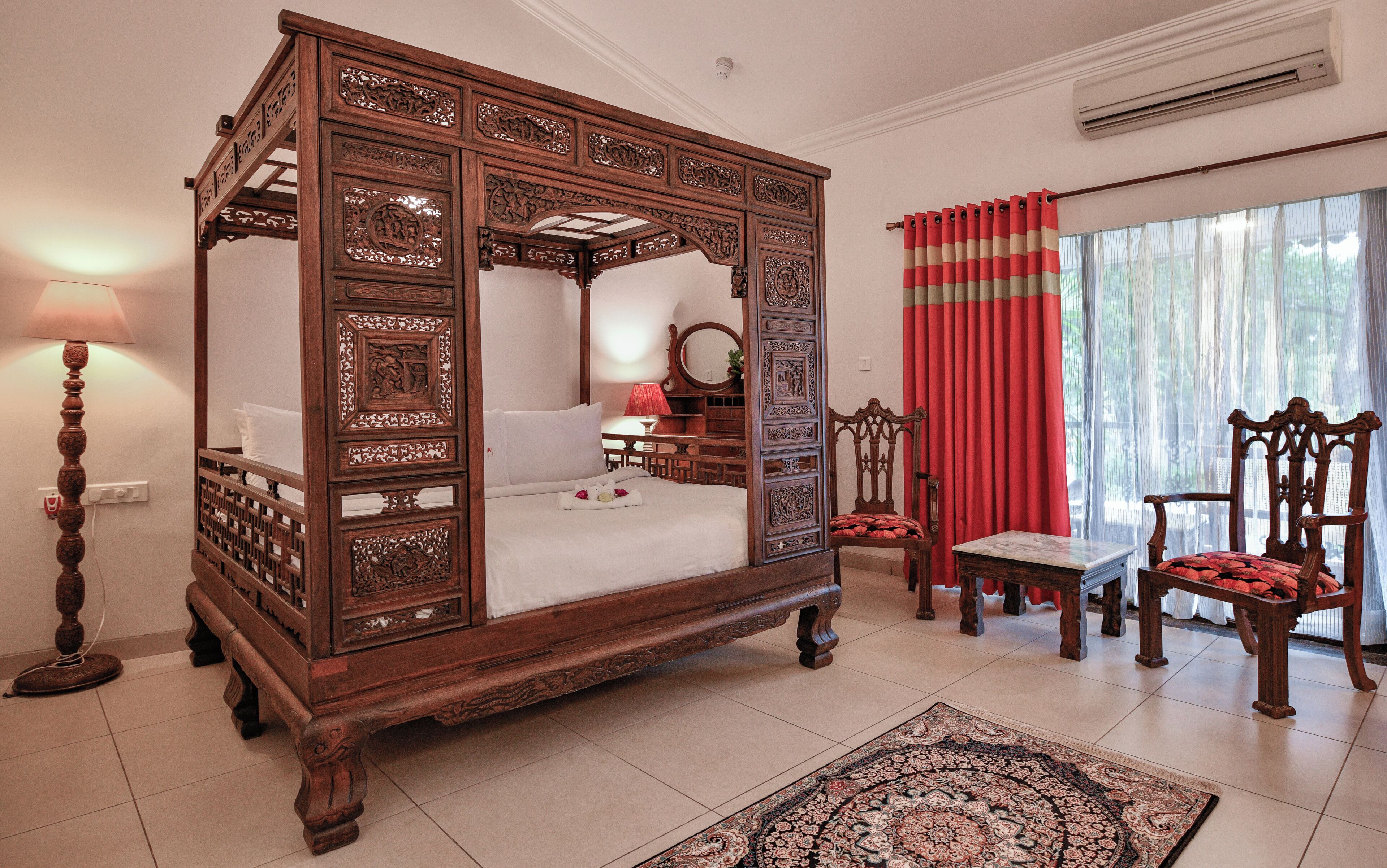 Una cama con dosel adornada con intrincados relieves, una mesita de noche con lámpara, un sillón con un cojín rojo y un ventanal con cortinas traslúcidas.