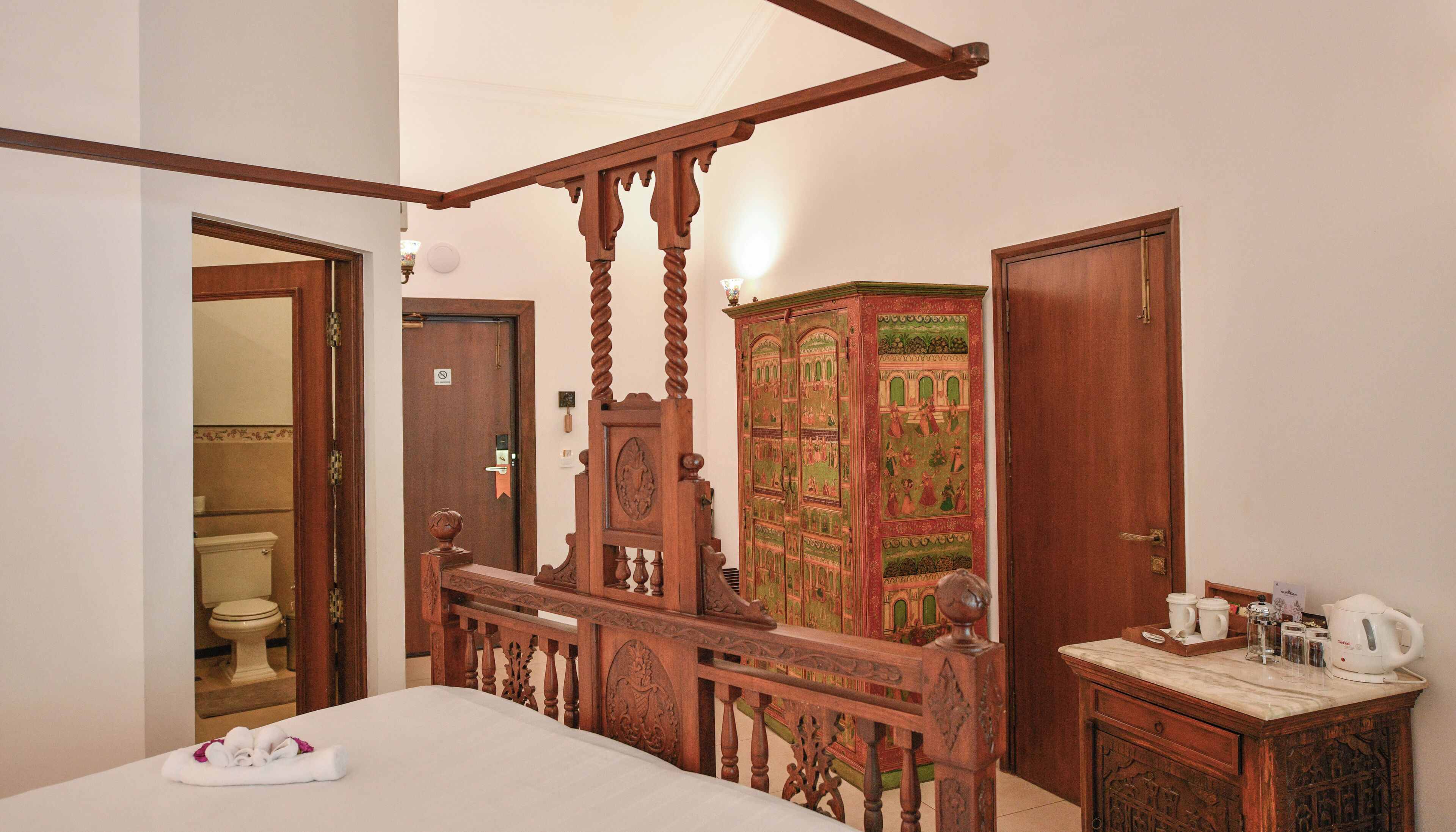 Una habitación con cama con dosel, un mueble de madera con diseños intrincados, un baño con inodoro y un rincón para preparar café.