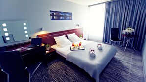 A hotel room with a bed, a desk with a laptop, a chair, a small table with a vase of flowers, and a large window with curtains.