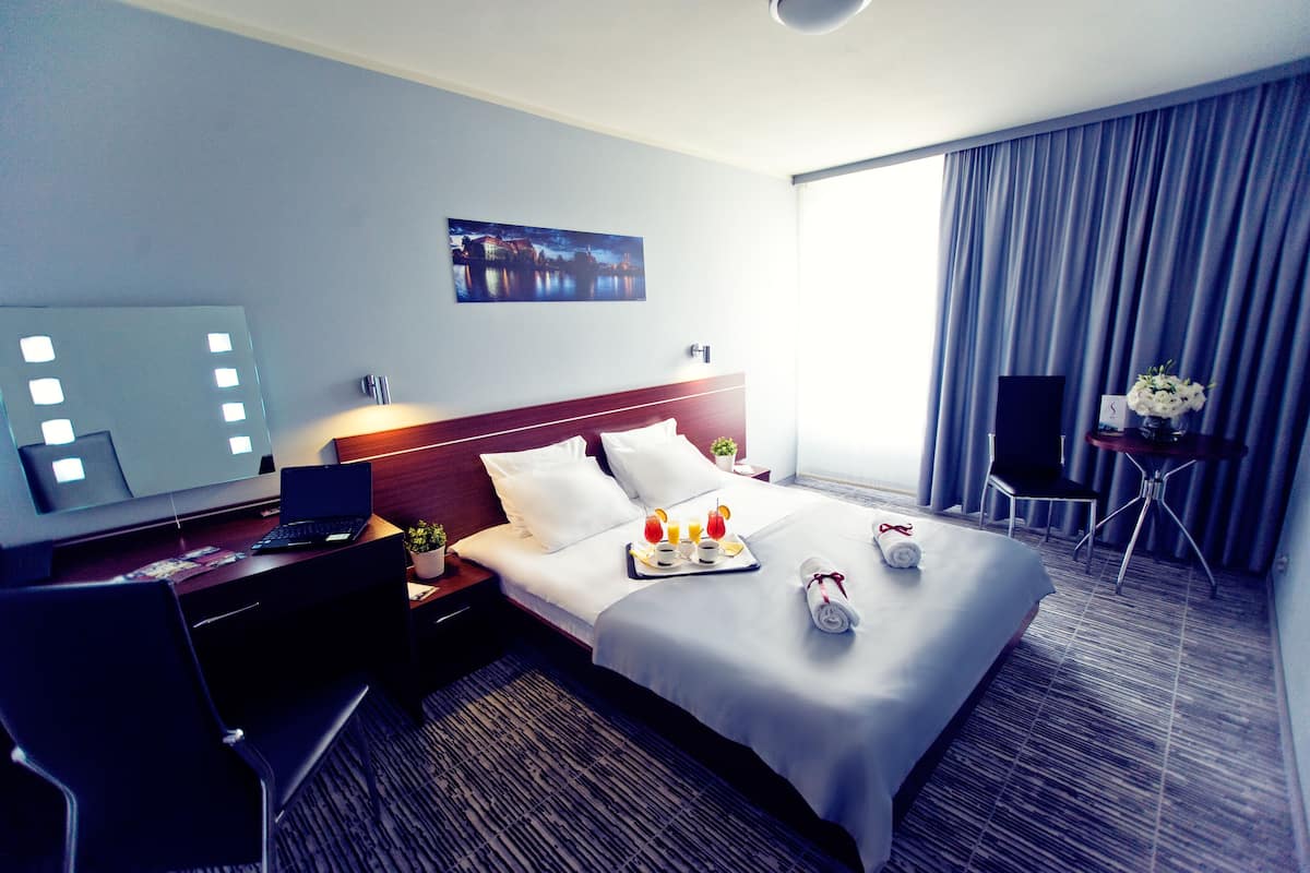 Ett hotellrum med en säng, ett skrivbord med en bärbar dator, en stol, ett litet bord med en vas med blommor och ett stort fönster med gardiner.