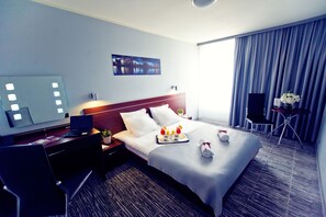 A hotel room with a bed, a desk with a laptop, a chair, a small table with a vase of flowers, and a large window with curtains.