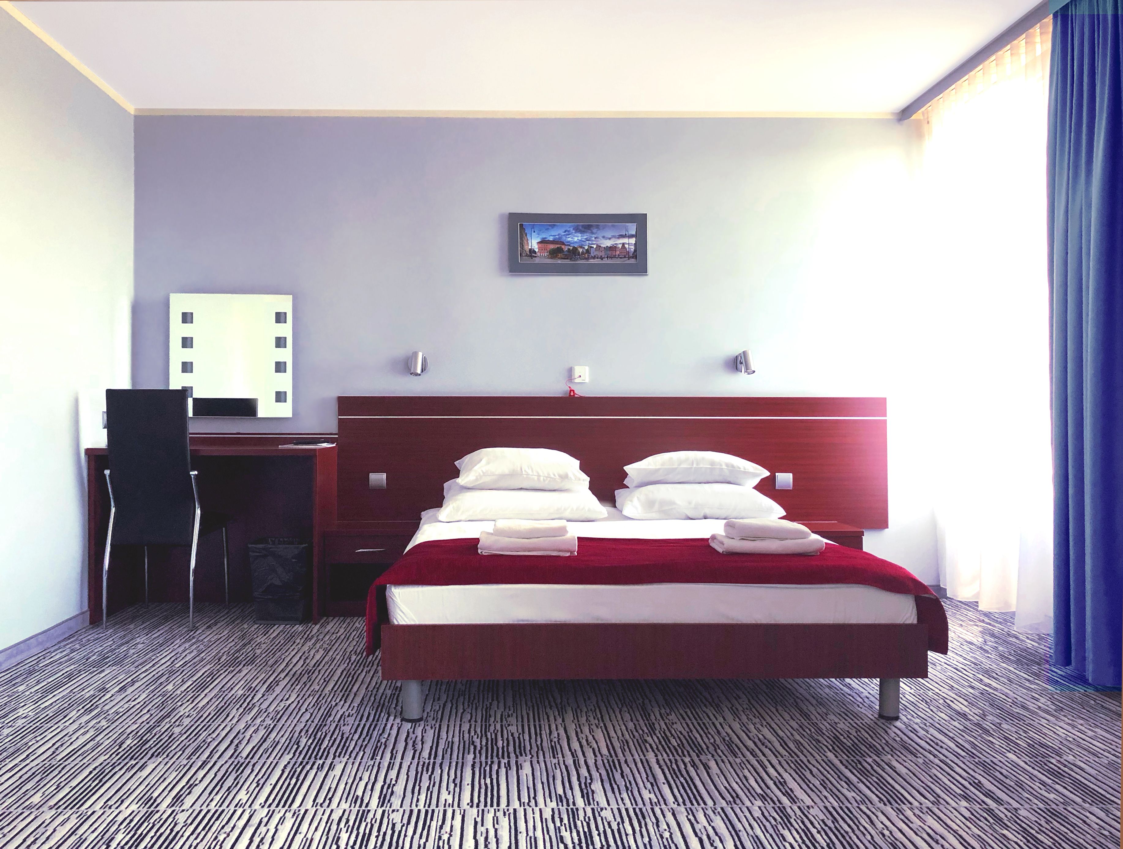 A hotel room with a bed, a desk, a chair, a wall-mounted TV, and a painting on the wall.