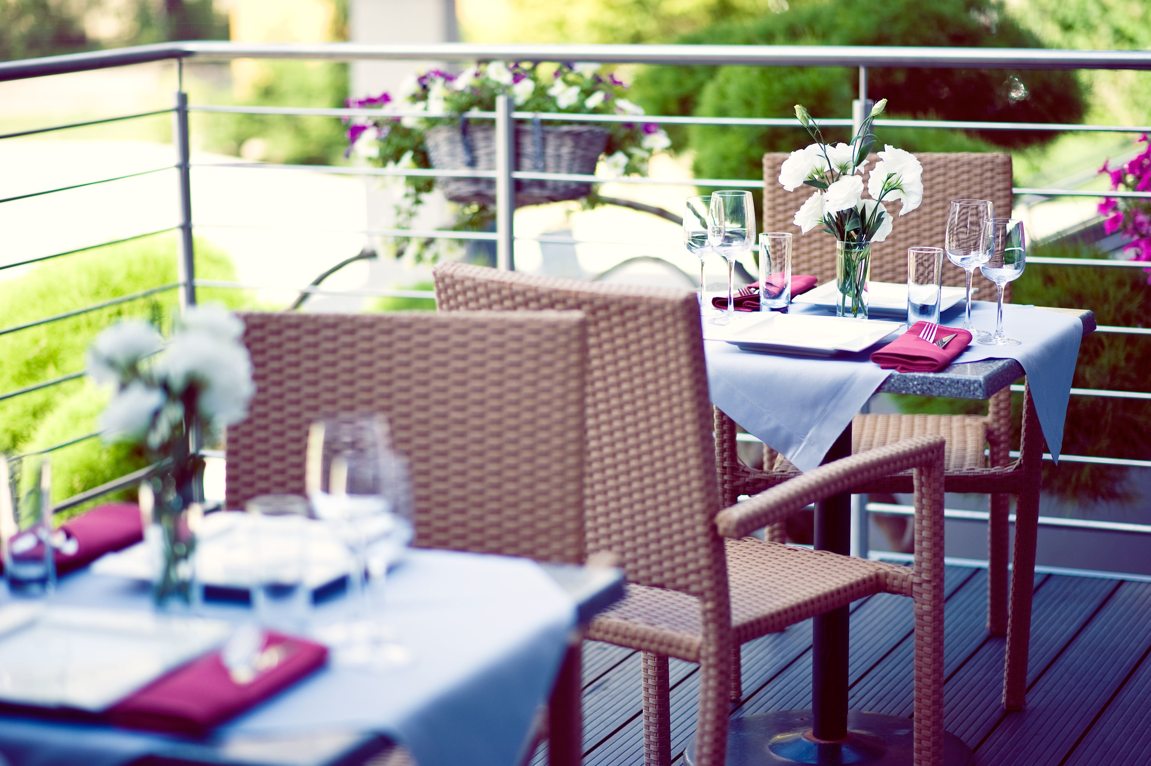 Un coin repas avec des chaises et des tables en osier, disposées pour deux personnes, chacune agrémentée d’une nappe blanche, d’un vase de fleurs et de verres à vin.