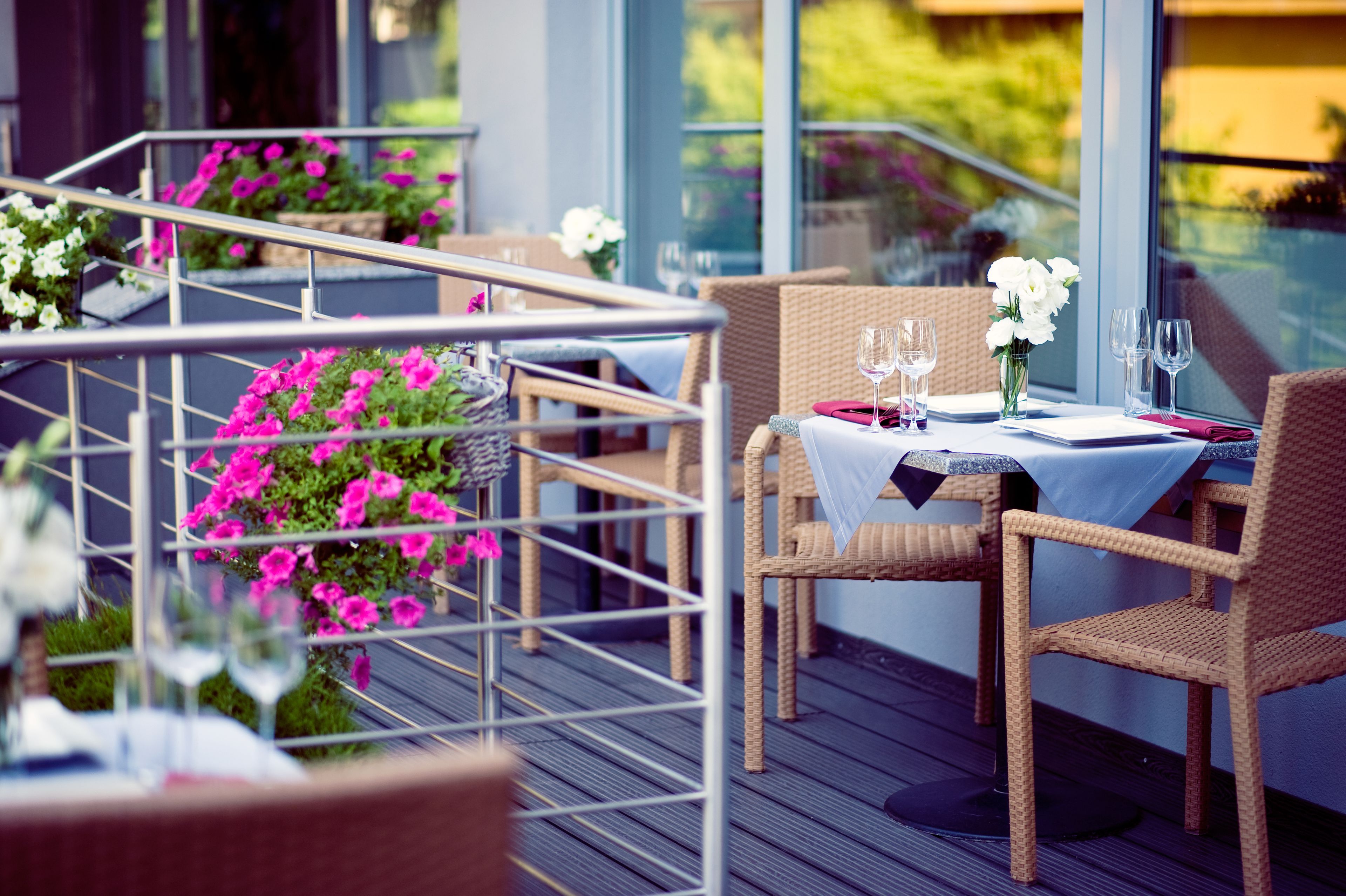 An outdoor dining area with wicker chairs and tables, a metal railing, and potted plants with pink flowers.
