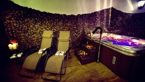 A spa area with a hot tub, two lounge chairs, candles, and a wall of pebbles.