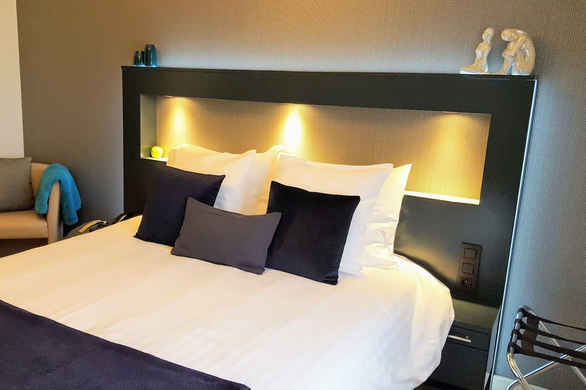 A neatly made bed with white and dark pillows, a headboard with built-in lighting, and a chair with a blanket.
