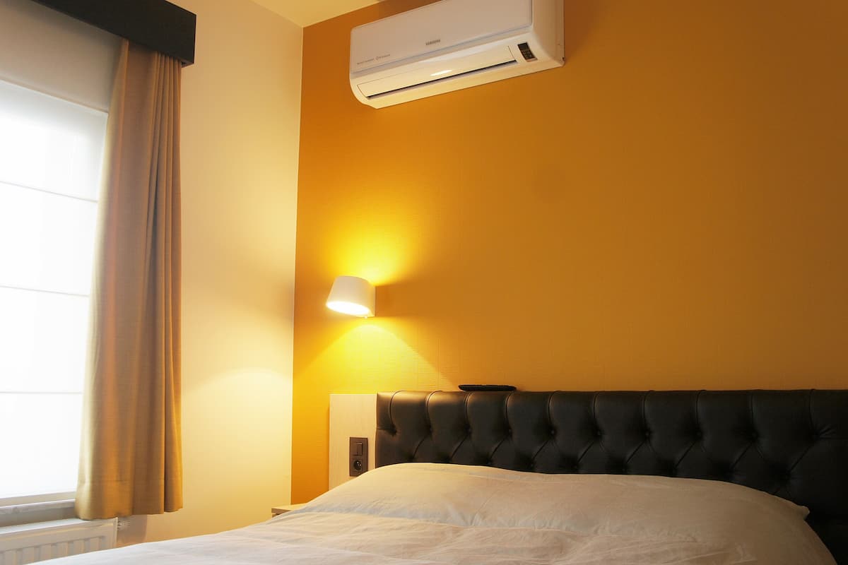 A hotel room with a bed, an air conditioner, a wall-mounted lamp, and a window with curtains.