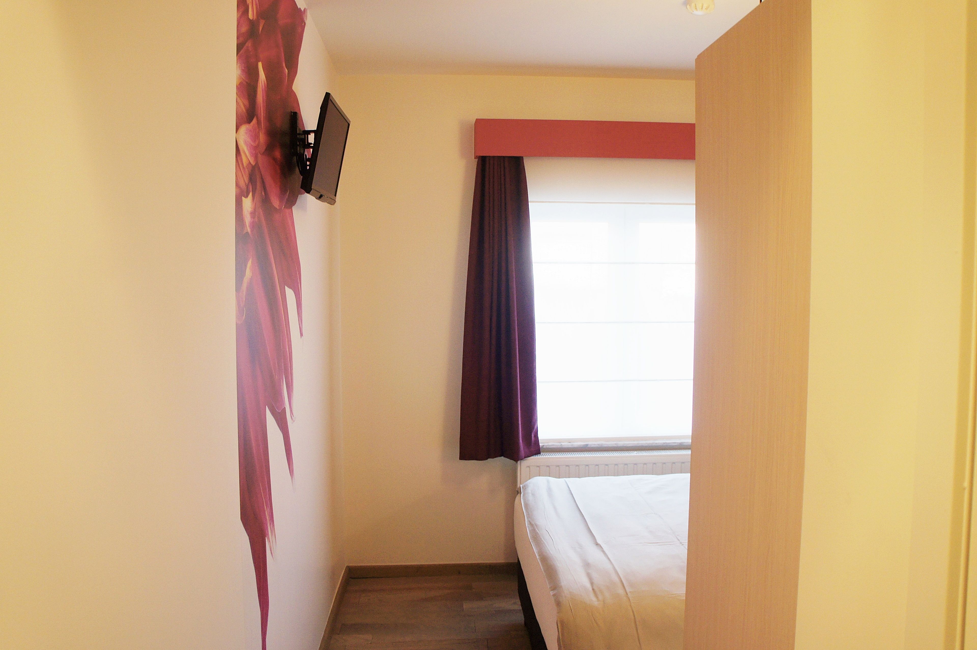 A hotel room with a bed, a wall-mounted TV, a window with a curtain, and a radiator.