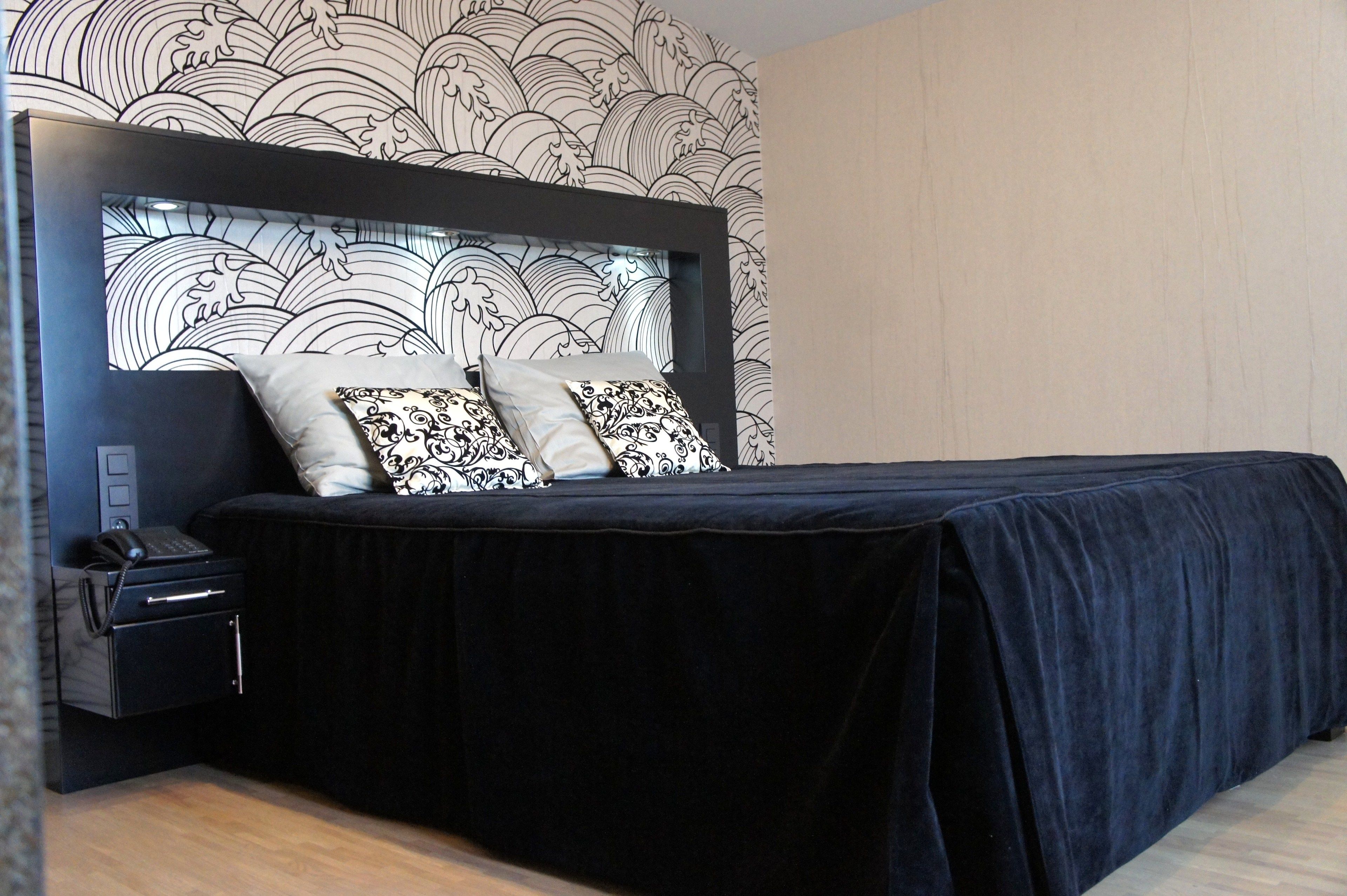 A modern bedroom with a dark headboard, two patterned pillows, a black bedspread, a bedside table with a phone, and a wall with a wave pattern.