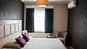 Une chambre d’hôtel avec un grand lit, une table de chevet, une chaise, une fenêtre avec des rideaux et un luminaire au plafond.