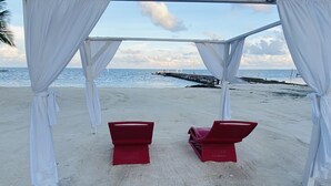 A beach setting with a white canopy structure and two red lounge chairs under it. - La Isla Resort (Caye Caulker)
