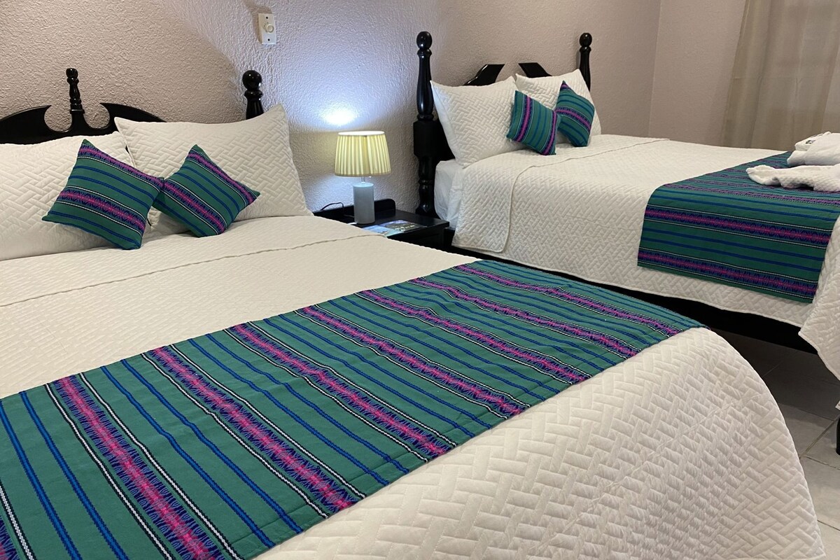 A hotel room with two beds, each with a white quilted bedspread and pillows. There is a lamp on a bedside table.