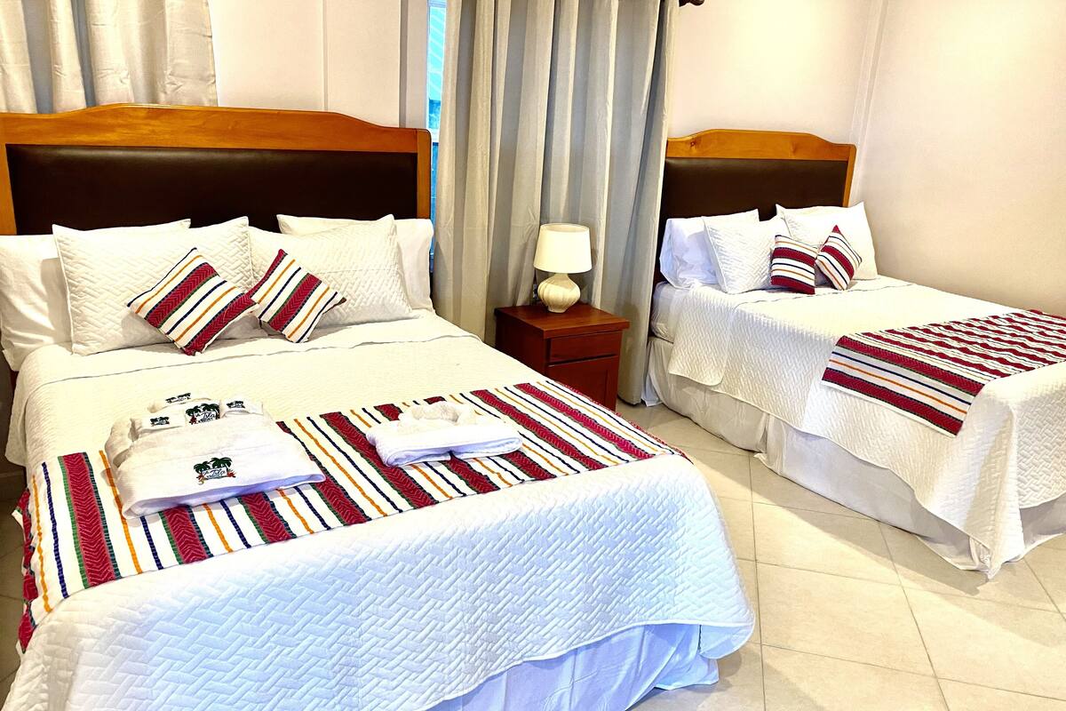 A hotel room with two beds, each with white bedding and striped pillows. There is a nightstand with a lamp between the beds.