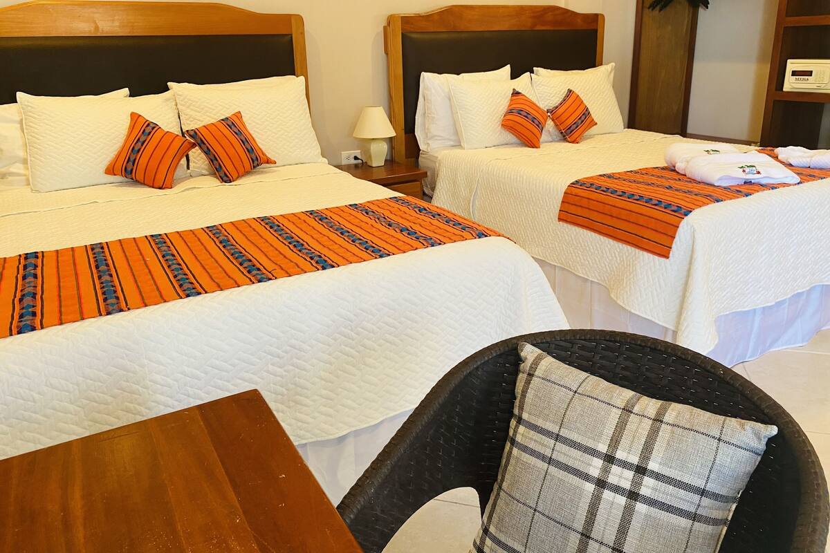 A hotel room with two beds, each with white bedding and orange-striped blankets. There is a wooden nightstand with a lamp, a black wicker chair, and a plaid pillow.