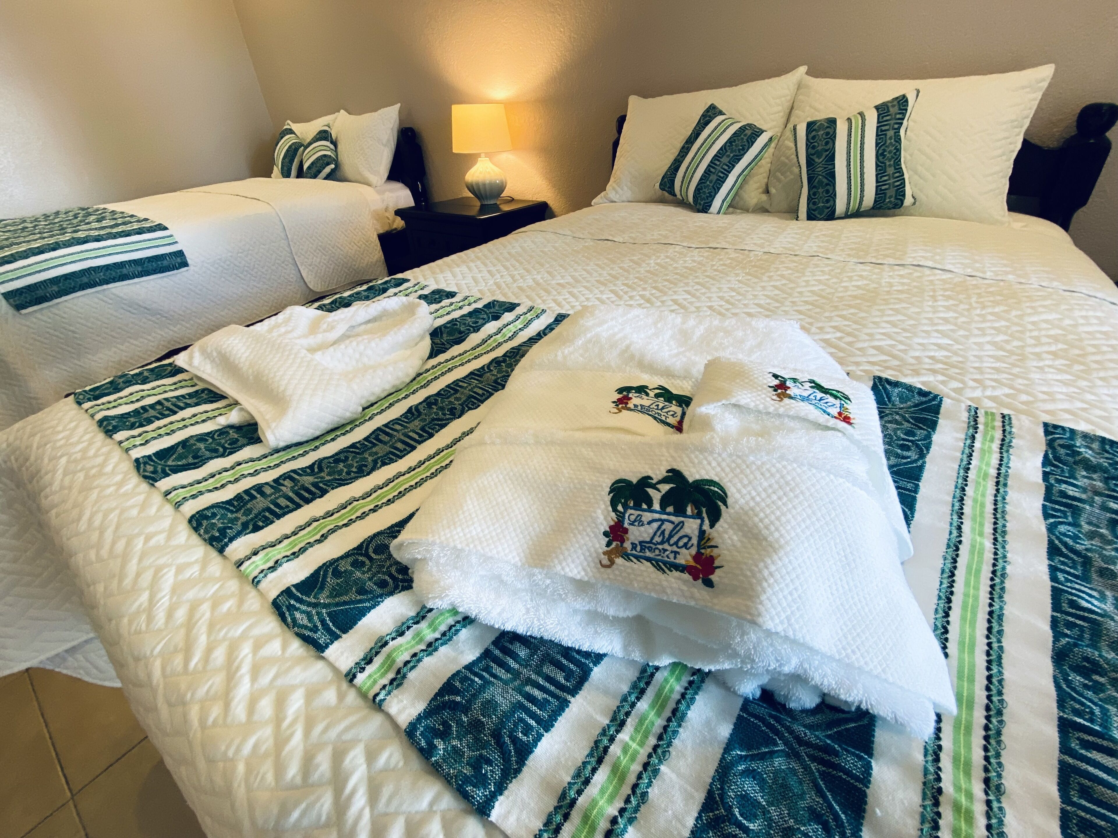 A hotel room with two beds, each with a white quilt and green and white striped blankets. There are towels with a palm tree design on the beds.