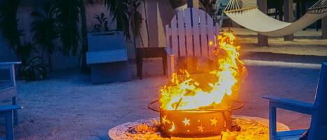 A fire pit with star decorations on a concrete base surrounded by chairs, set on a sandy surface with a hammock and chairs in the background.