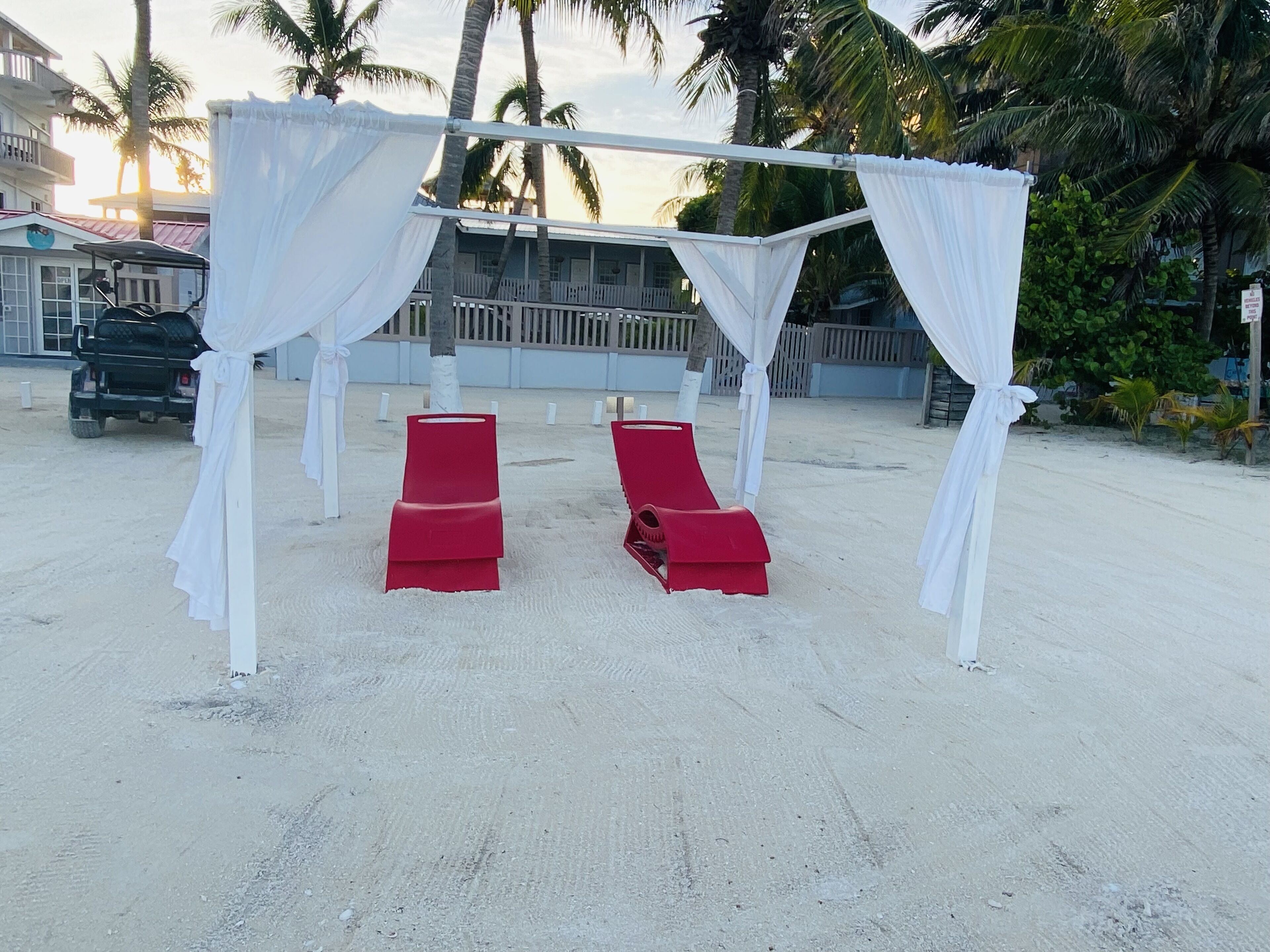 Un paisaje playero con una estructura tipo sombrilla, dos sillones reclinables rojos y un carrito de golf.