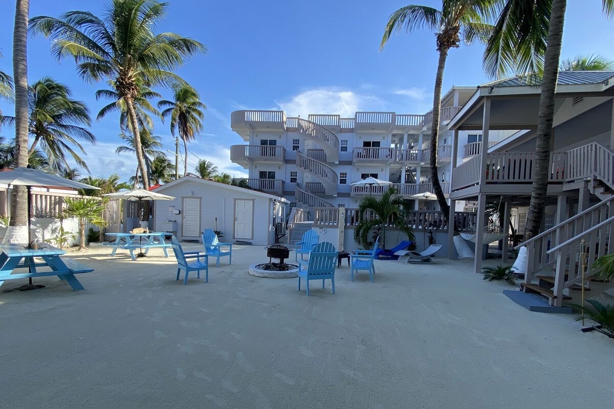 Hotel frente a la playa con varios balcones, mobiliario exterior y palmeras.