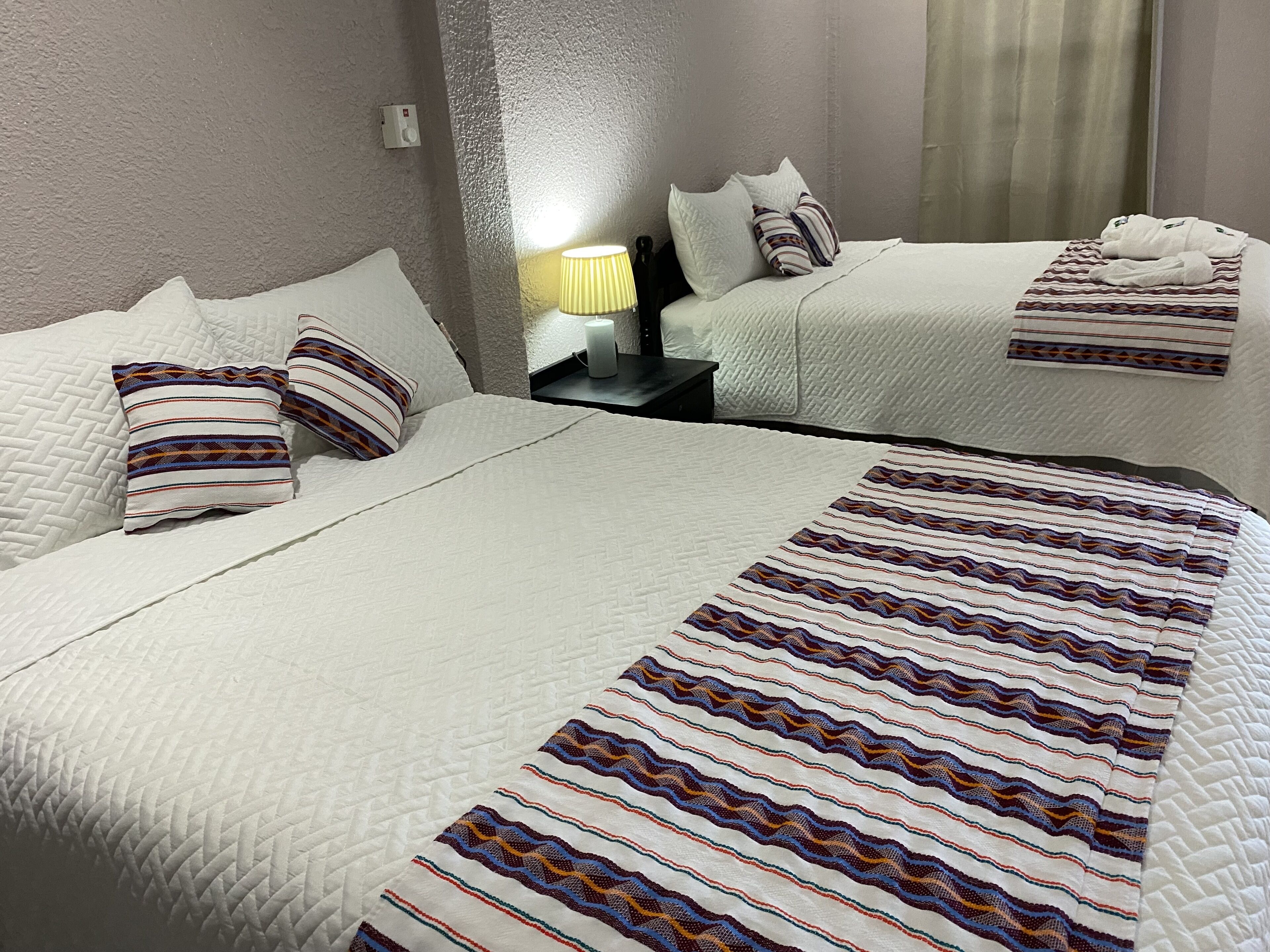 A hotel room with two beds, each with white bedding and striped pillows. There is a nightstand with a lamp between the beds.