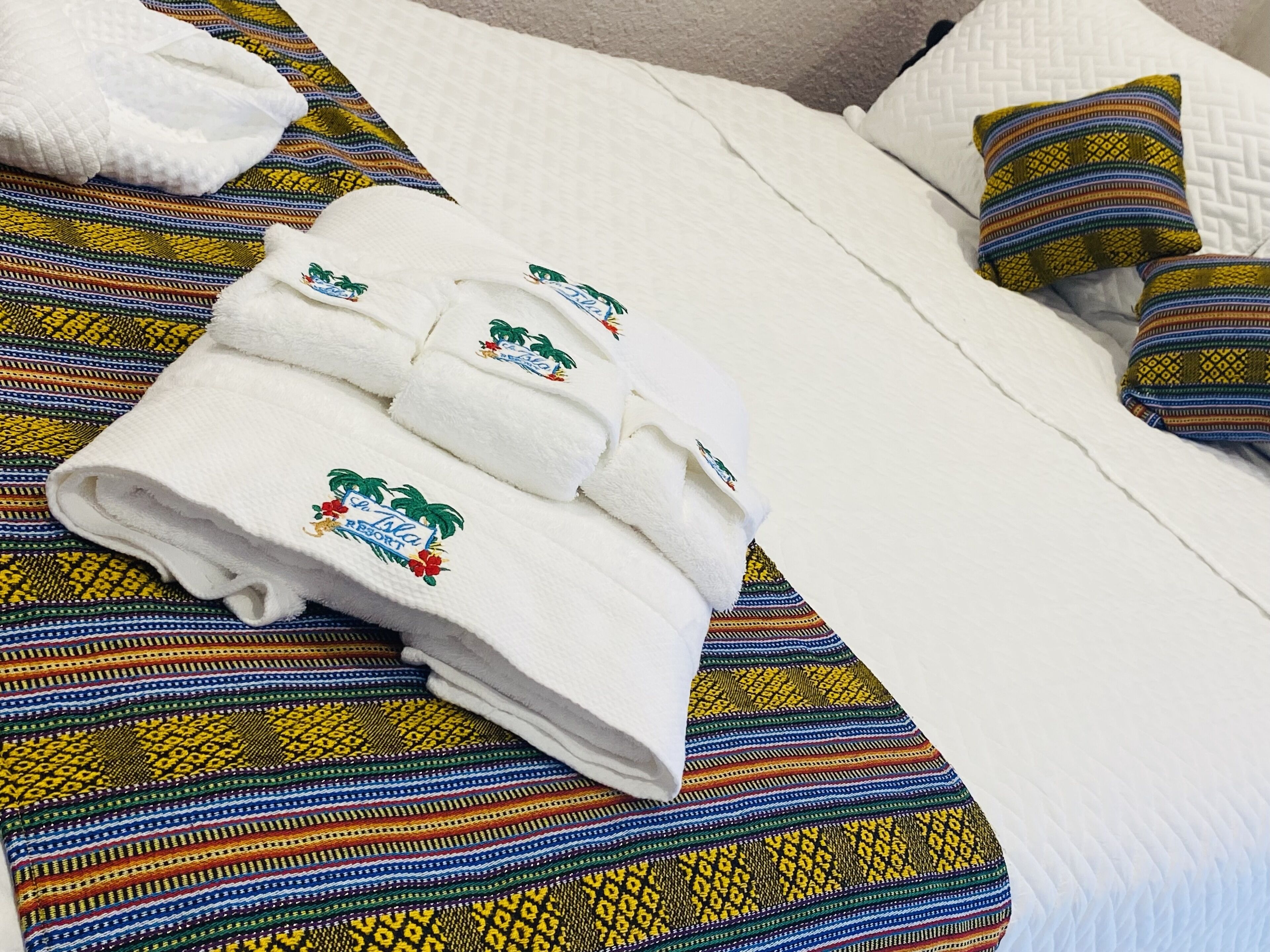 Una cama bien tendida con un colorido edredón a rayas, una almohada blanca y una toalla blanca doblada con motivos tropicales.