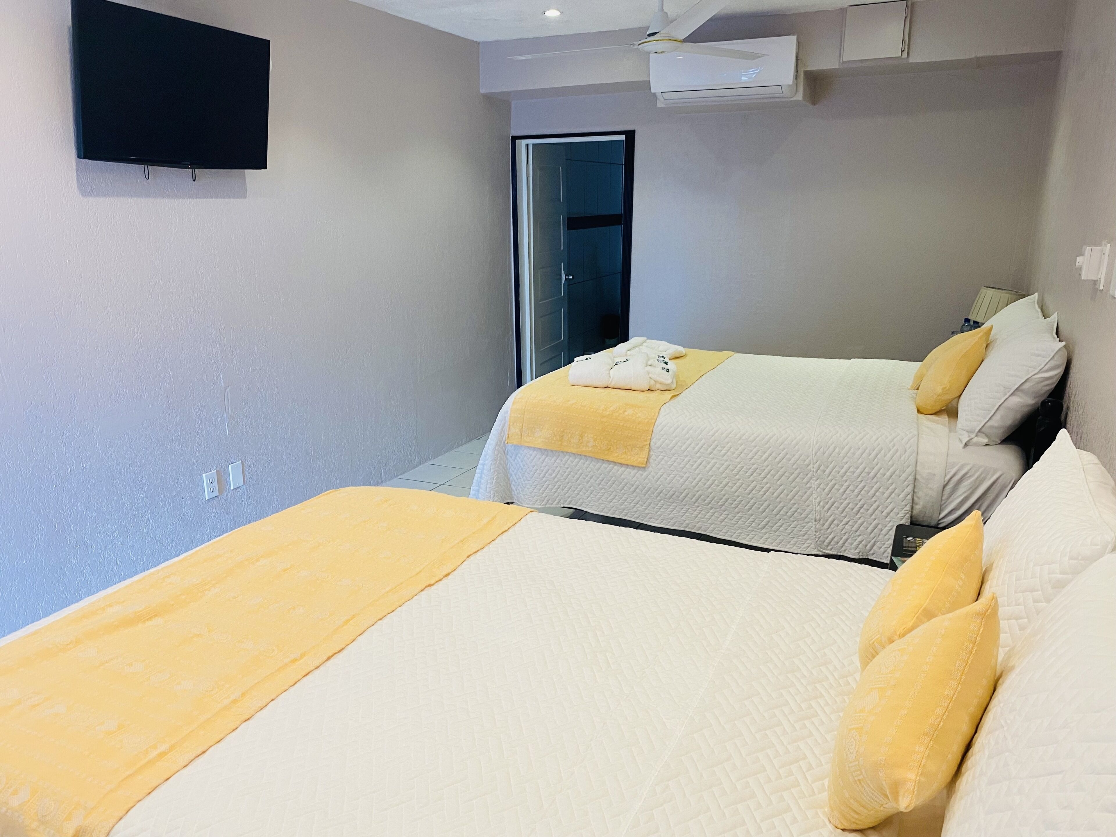 Ein Hotelzimmer mit zwei Betten, jeweils mit einer gelben Decke und Kissen. An der Wand ist ein Flachbildfernseher befestigt und über den Betten befindet sich eine Klimaanlage.