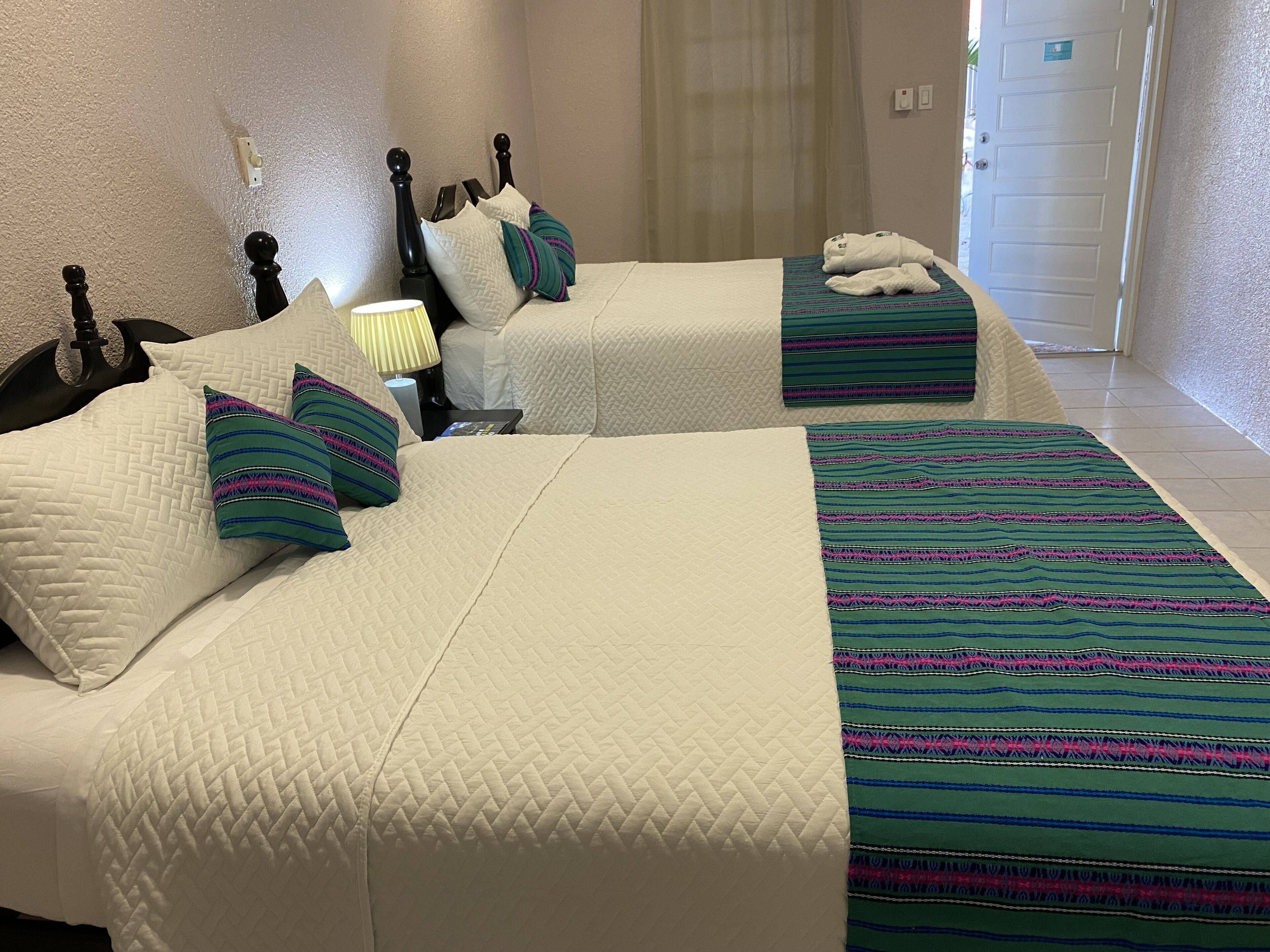 Habitación de hotel con dos camas, cada una con ropa blanca y mantas a rayas. Hay una mesita de noche con una lámpara y una pequeña mesa con un libro.