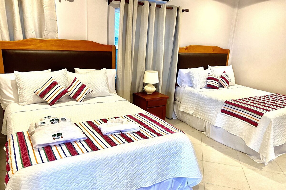 A hotel room with two beds, each with white bedding and striped pillows, a nightstand with a lamp, and a window with curtains.