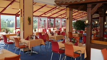 Ein Restaurant mit mehreren Tischen und Stühlen, großen Fenstern und einer Holzbalkendecke.
