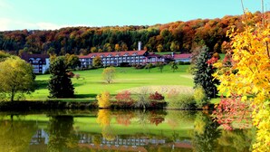 A hotel with a large building, a pond in the foreground, and autumn foliage in the background. - Hessen Hotelpark Hohenroda (Hohenroda)