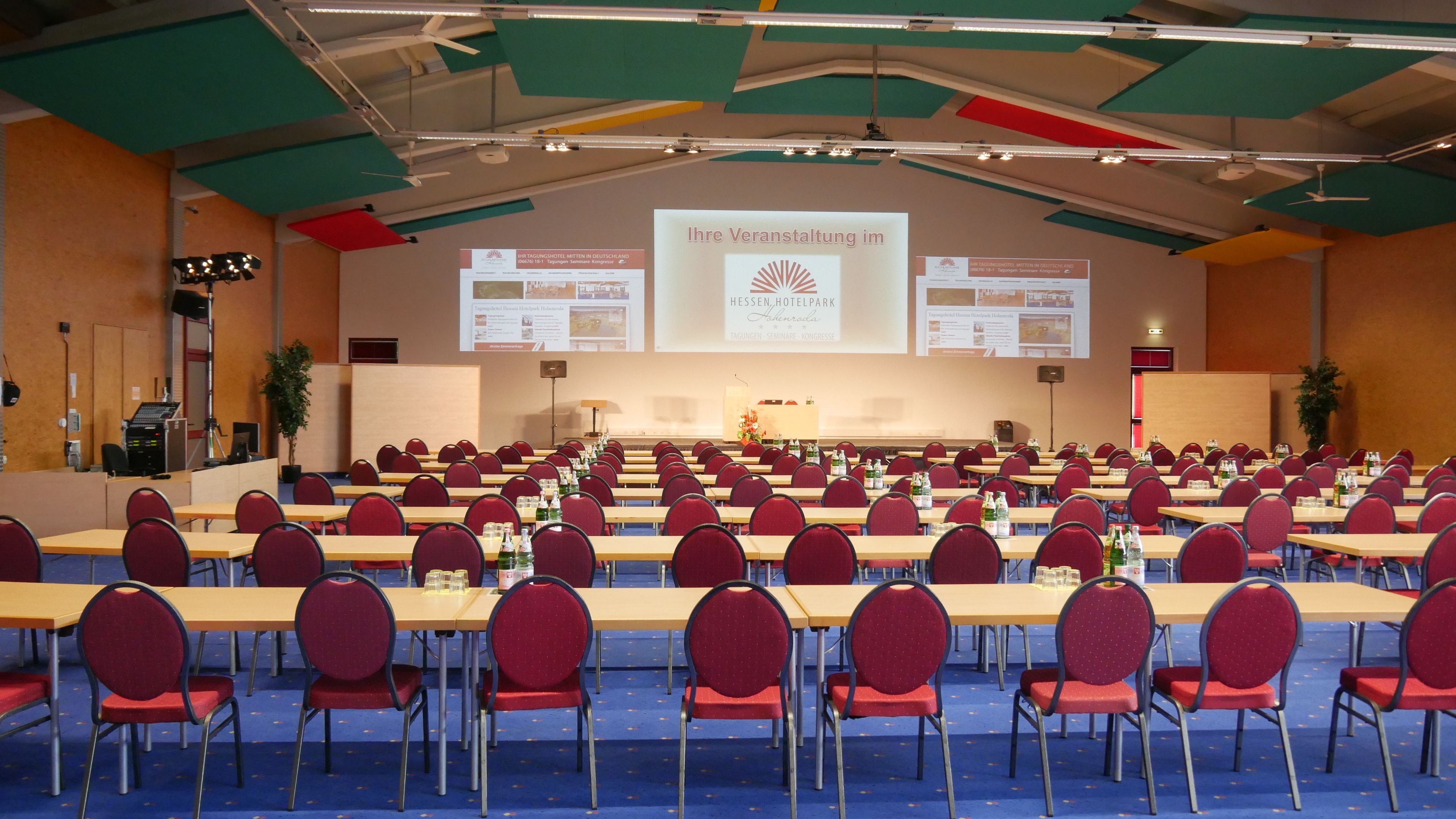 A conference room with rows of tables and chairs, a stage with a presentation screen, and a ceiling with colorful panels.