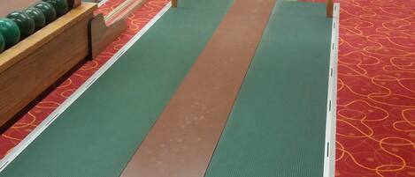 Una bolera con pistas verde y marrón, un soporte de madera para bolas de boliche y un piso cubierto de alfombra roja.