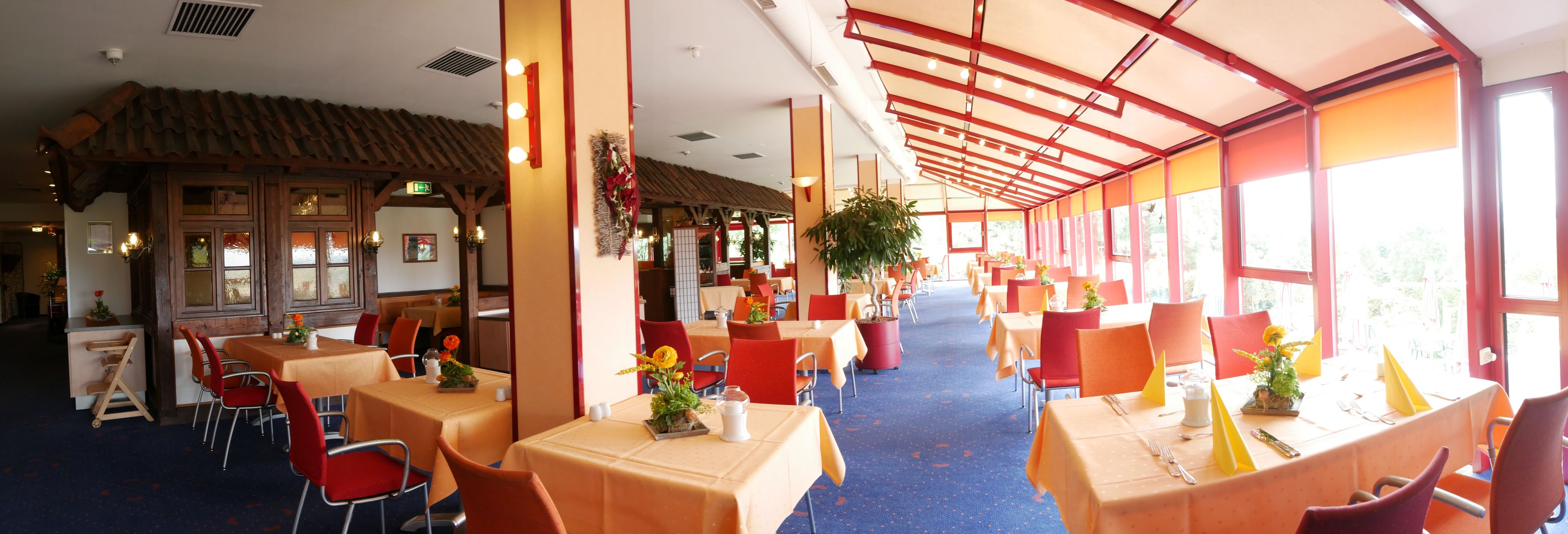 A restaurant with tables and chairs, a ceiling with a red beam structure, and large windows allowing natural light.