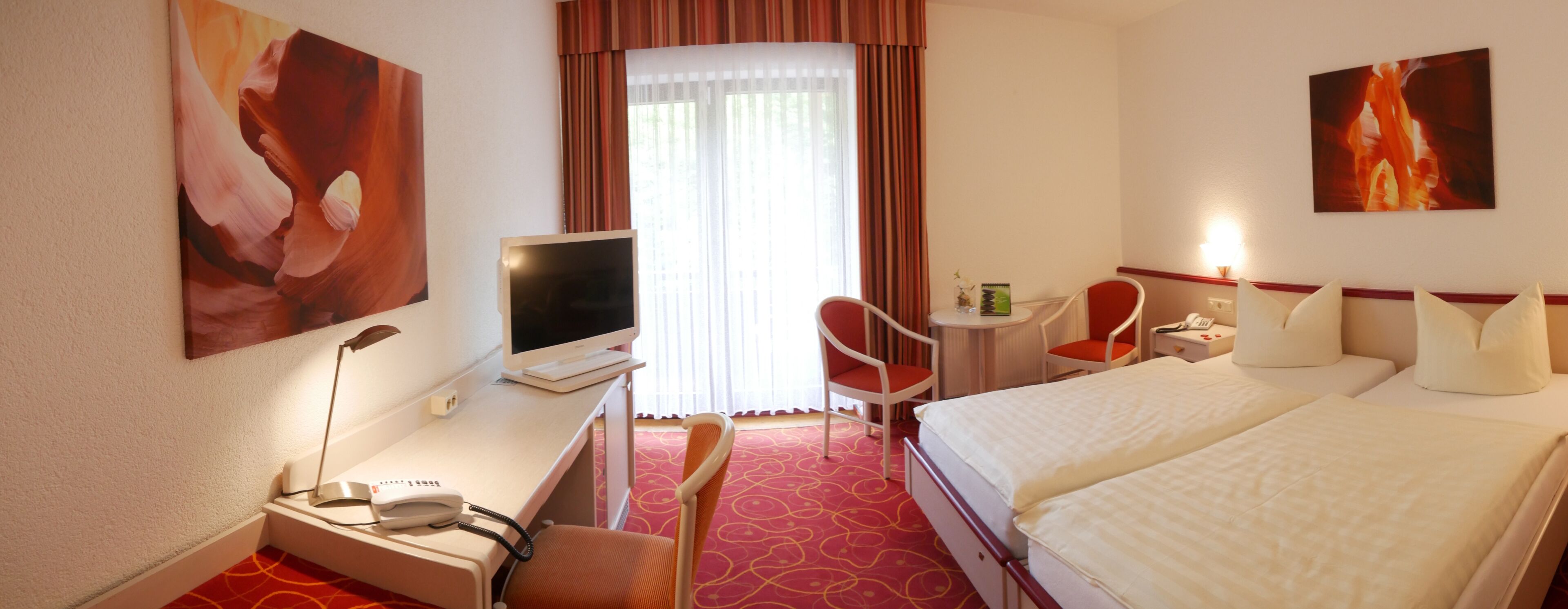 A hotel room with a bed, desk, chair, TV, and artwork on the walls.