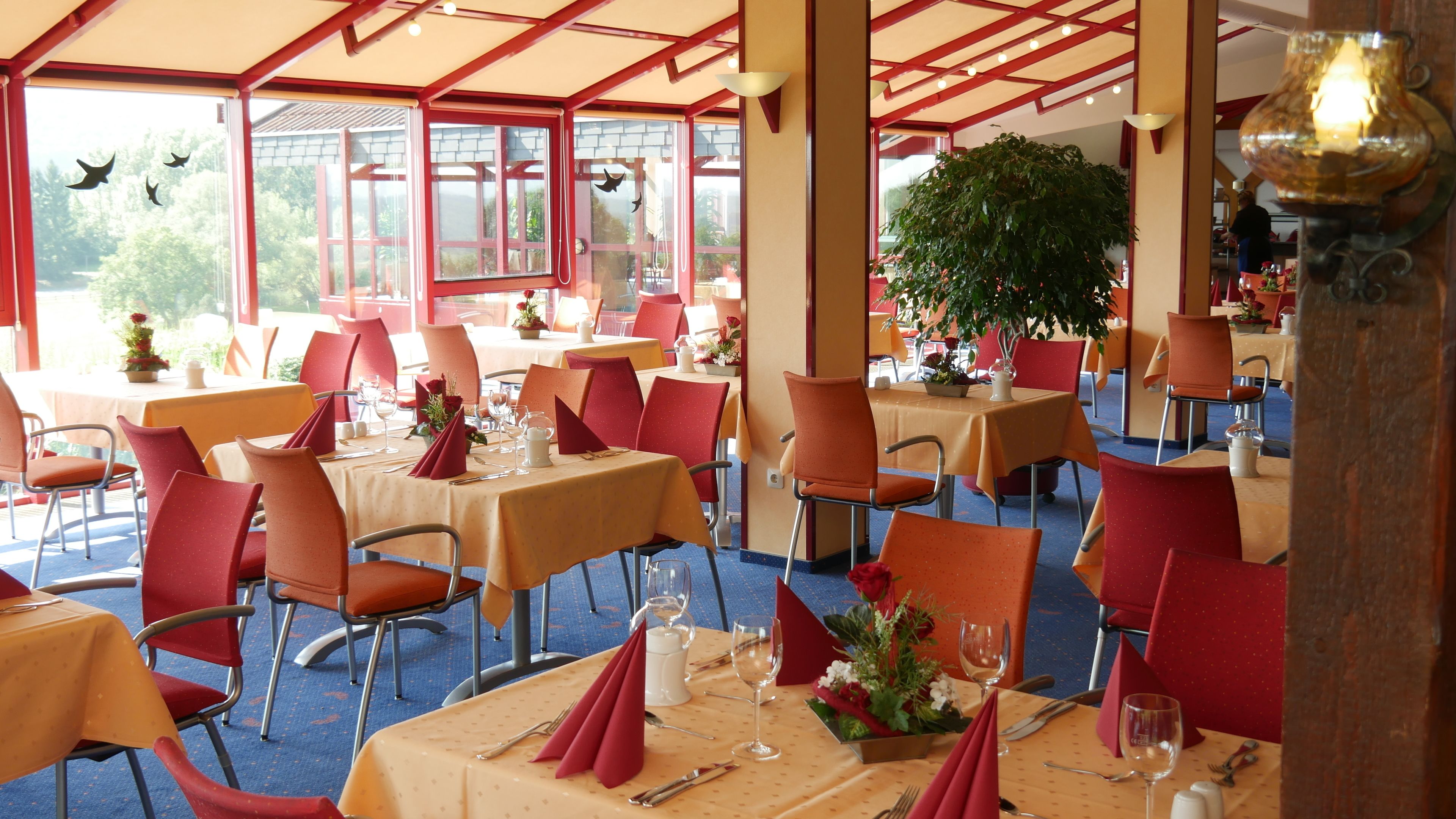 A restaurant with tables set for dining, red chairs, and a large potted plant in the center.