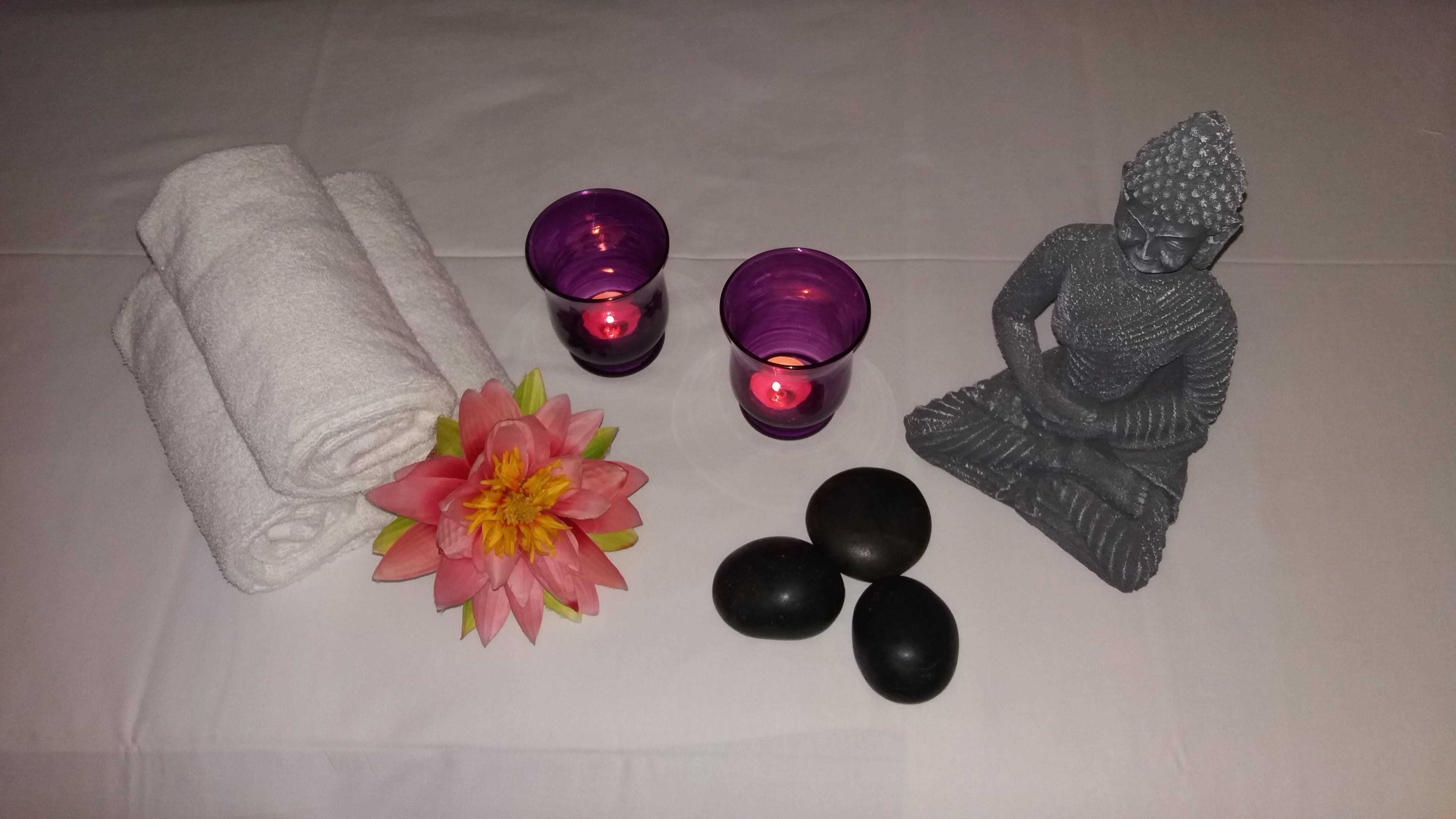 Una toalla blanca, dos velas moradas, una flor rosa, una estatua negra y tres piedras negras sobre una superficie blanca.