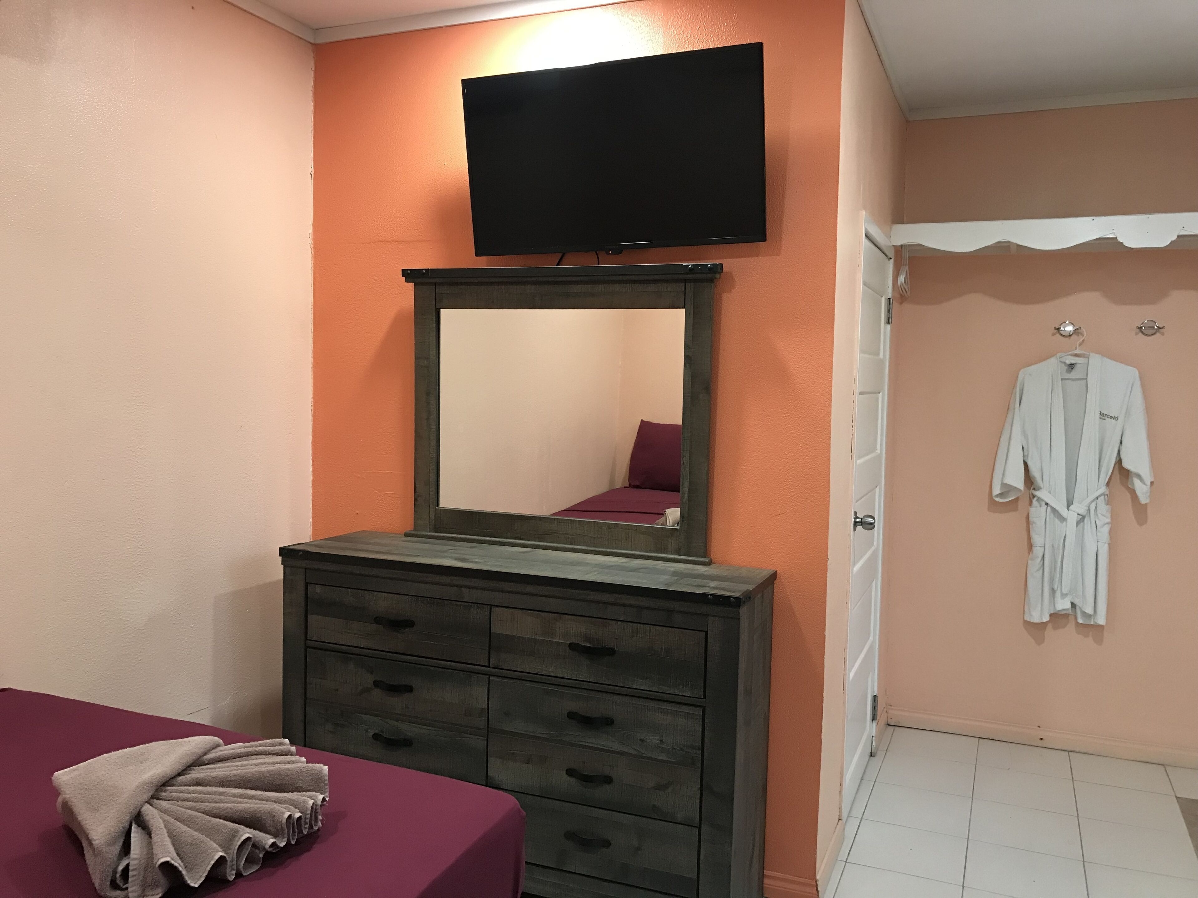 A room with a bed, a dresser, a television mounted above a mirror, and a robe hanging on a door.