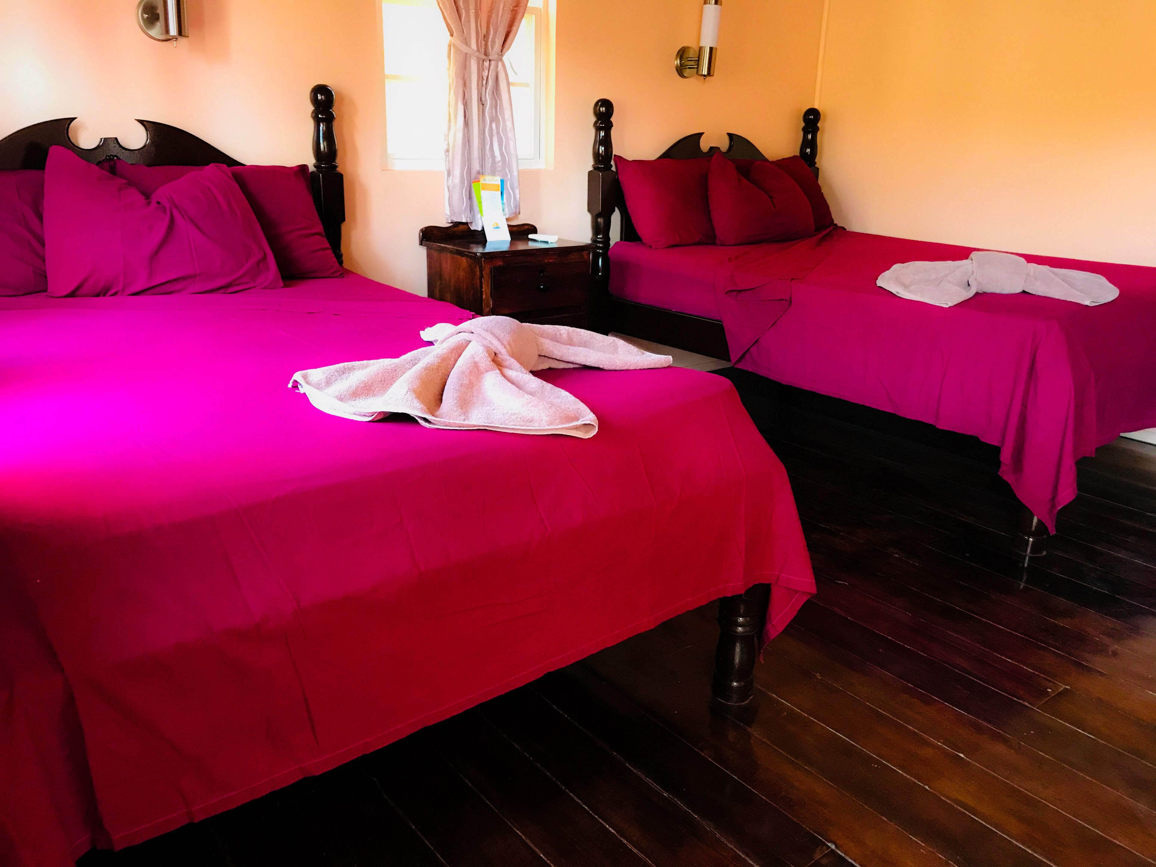 Camera d'albergo con due letti, entrambi con copriletto e cuscini rossi, pavimento in legno e un comodino con una lampada e una scatola.
