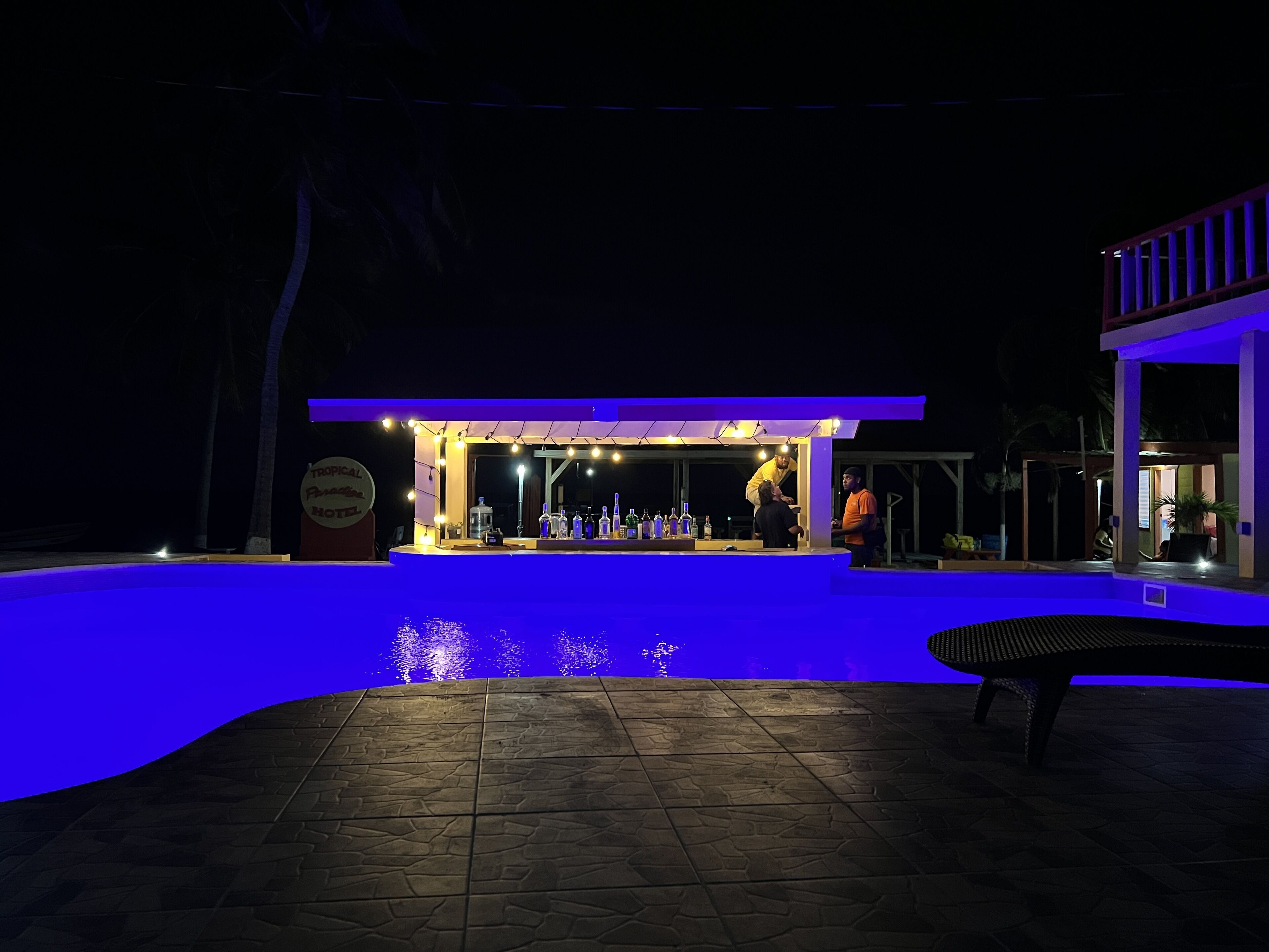 A nighttime bar area with a pool, illuminated by blue lighting, and a sign indicating a tropical theme.