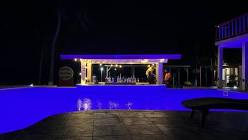 A nighttime bar area with a pool, illuminated by blue lighting, and a sign indicating a tropical theme.