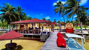 Un muelle de madera que lleva a un edificio con techo rojo, un cartel que indica Tropical Paradise Hotel, y una lancha roja amarrada cerca.