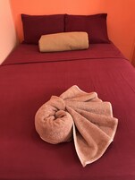 Una cama con una colcha roja, una almohada color beige y una toalla marrón doblada encima.