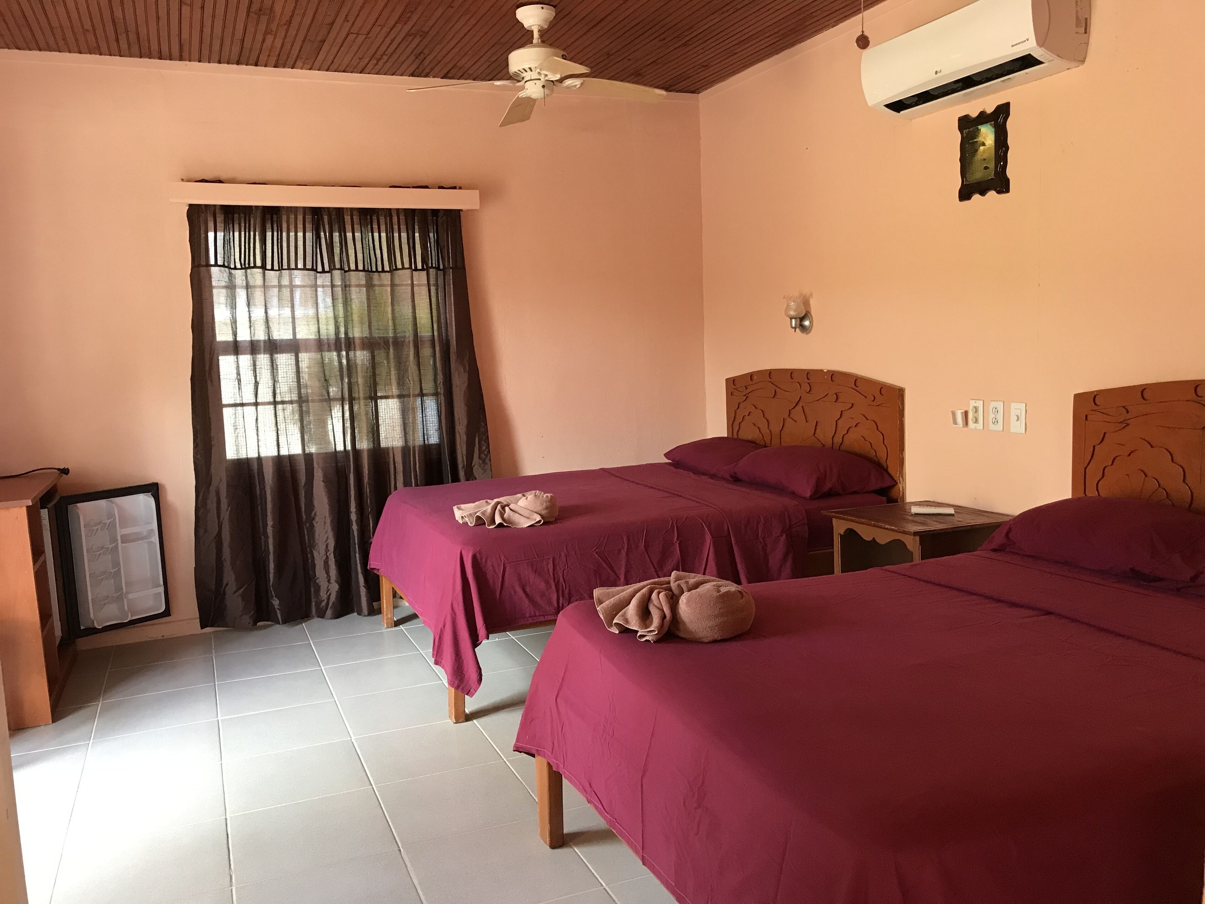 Une chambre avec deux lits, un ventilateur de plafond, un climatiseur, une fenêtre avec des rideaux et une petite table avec une lampe.