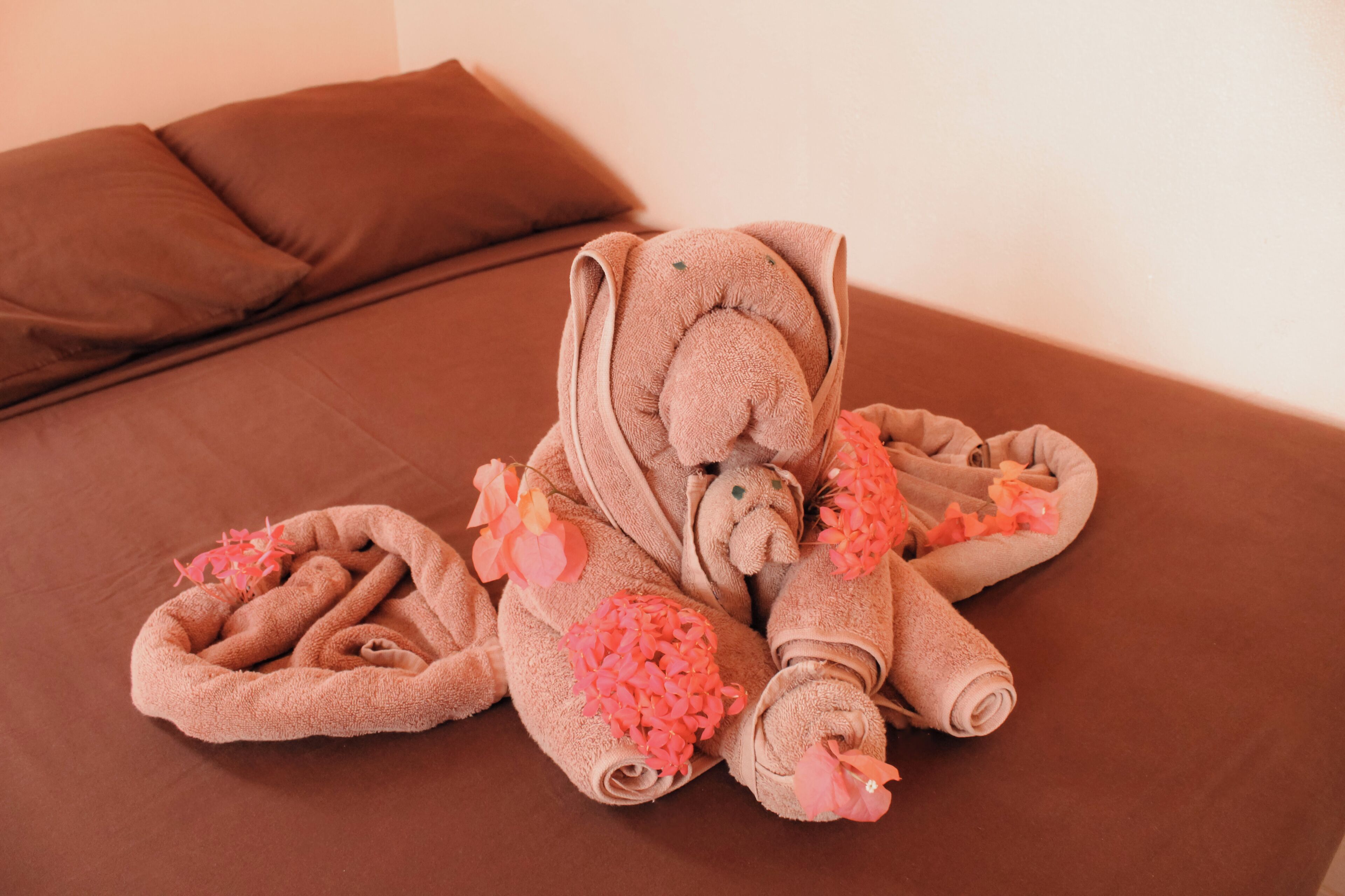 A bed with a brown cover, a brown pillow, and a towel shaped like an elephant with pink flowers.
