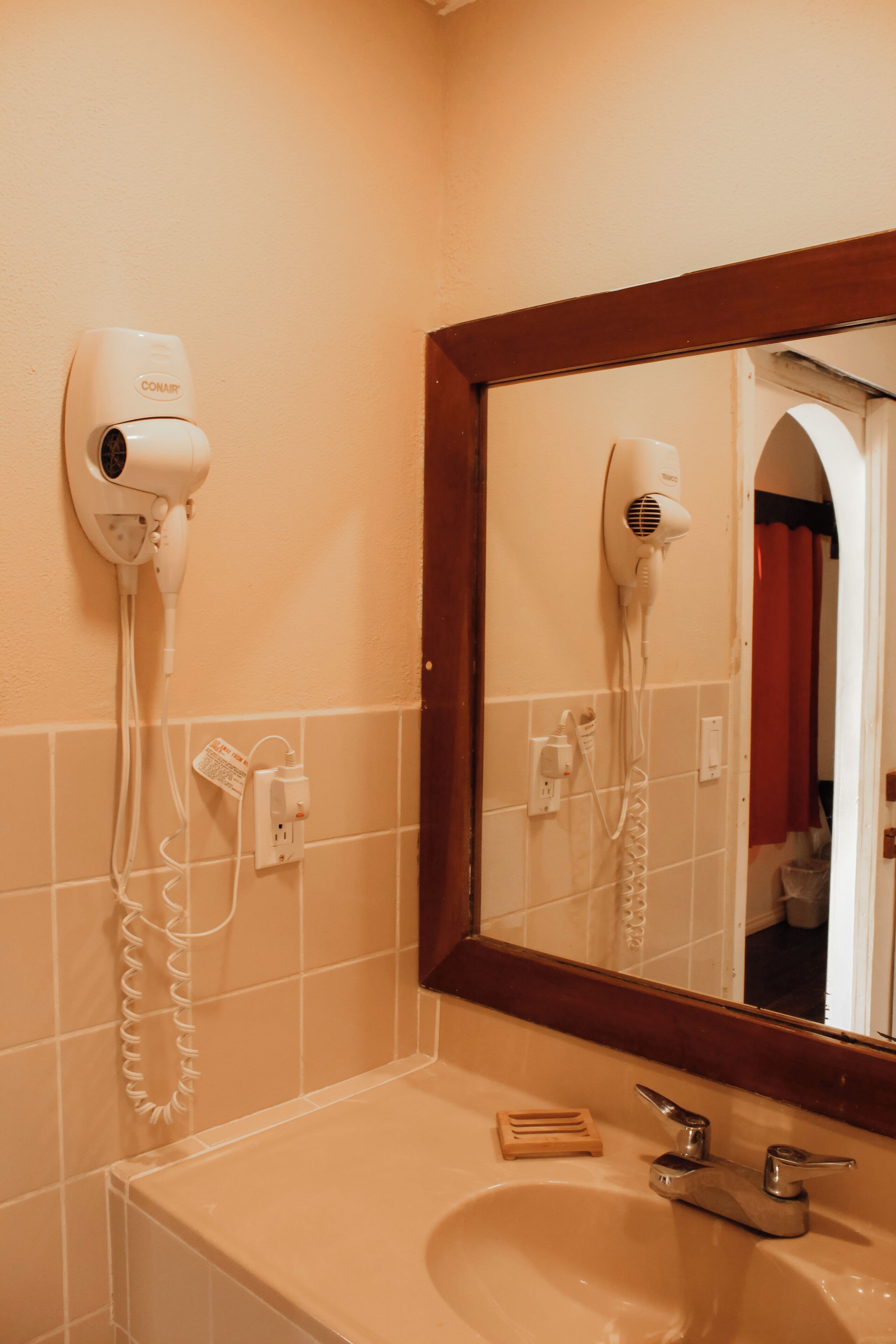 Een badkamer met een haardroger aan de muur, een wastafel en een spiegel die de ruimte weerspiegelt.