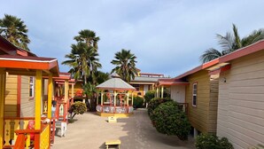 Un hilera de cabañas coloridas con un pabellón central, rodeadas de palmeras y suelo arenoso.